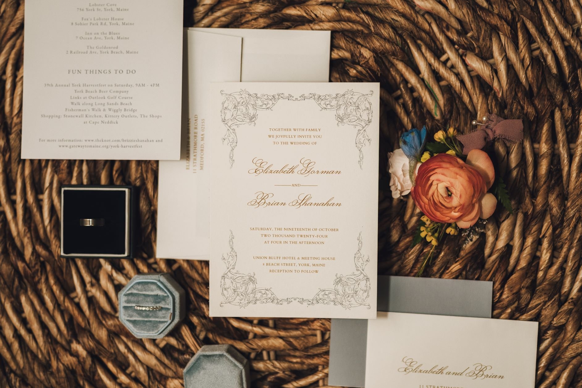 Wedding invitation set with floral arrangement and rings on woven background.