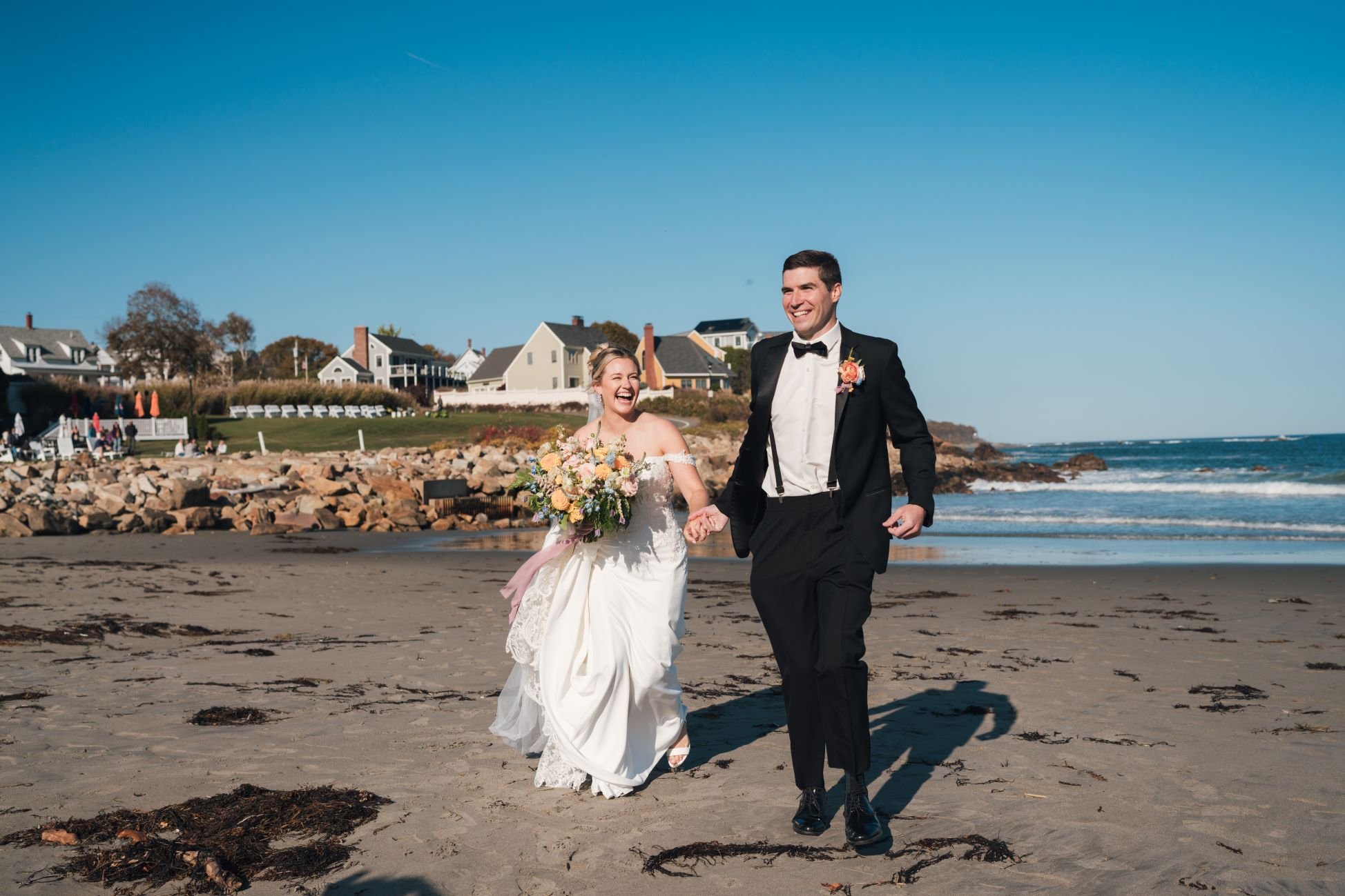 Bride and groom walking on a sandy beach, bride holding a bouquet, ocean waves in the background, clear blue sky.