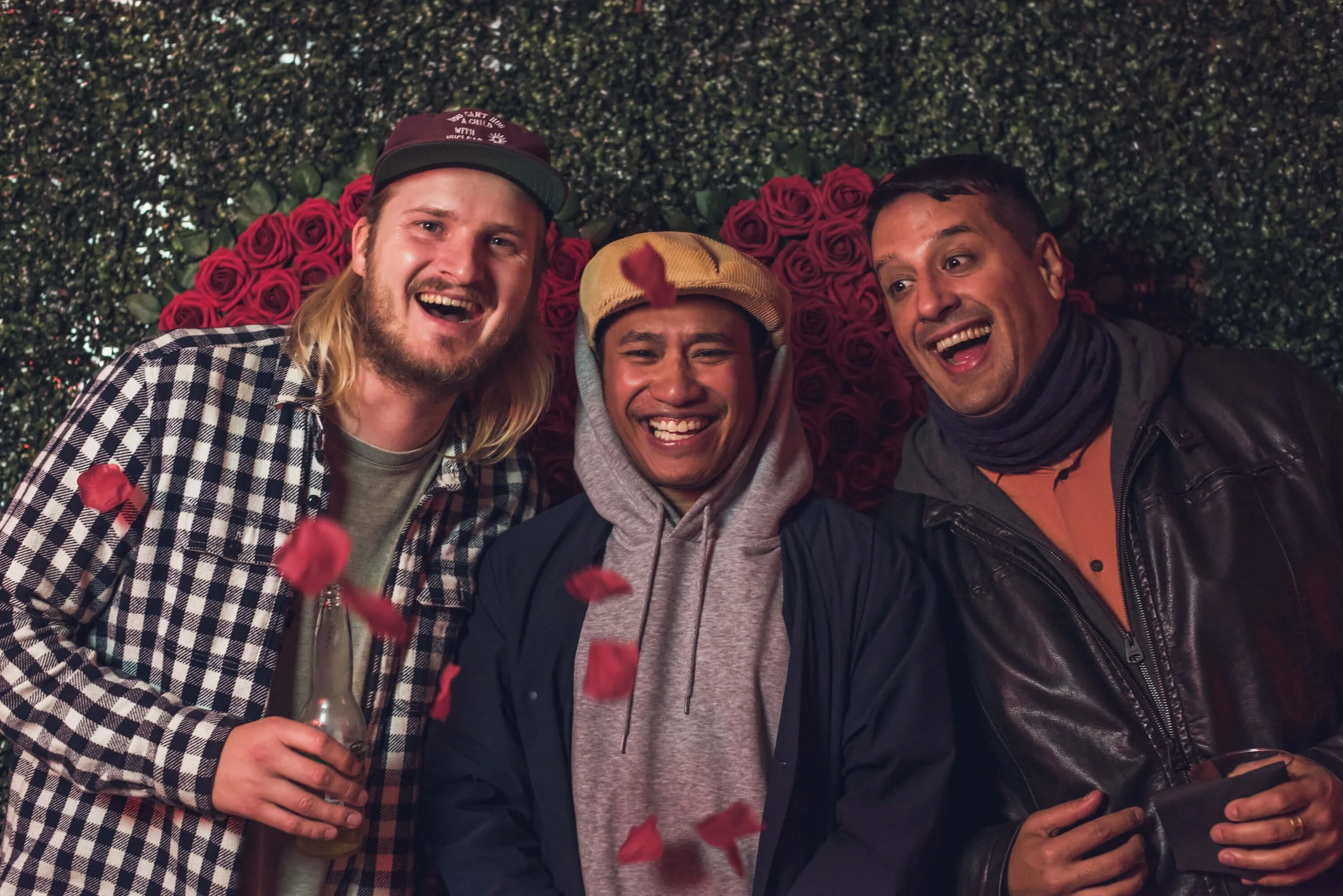 Three smiling men celebrating surrounded by falling rose petals with a backdrop of roses and greenery.