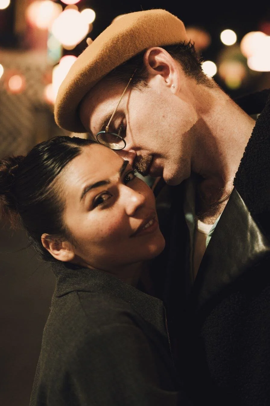 A young woman with dark hair tied back, smiling with her head tilted slightly, and a man with glasses, a beige hat, and a dark jacket are close together with their faces touching at night, with blurred colorful lights in the background.