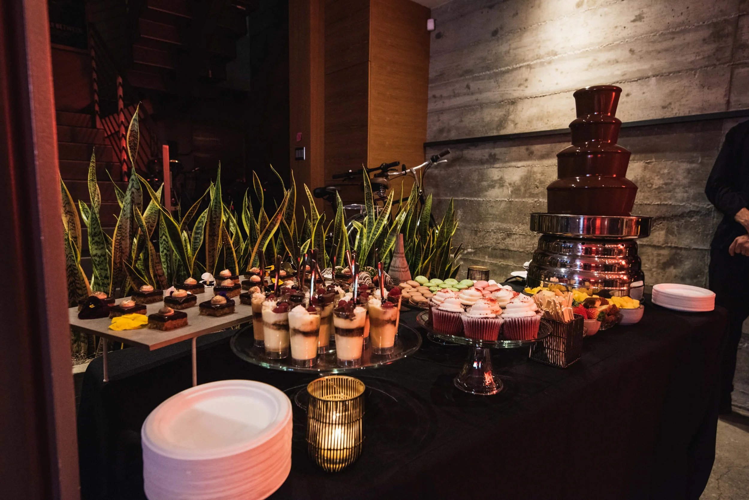 Dessert table with chocolate fountain, cupcakes, parfait glasses, macarons, and assorted treats at an indoor event.