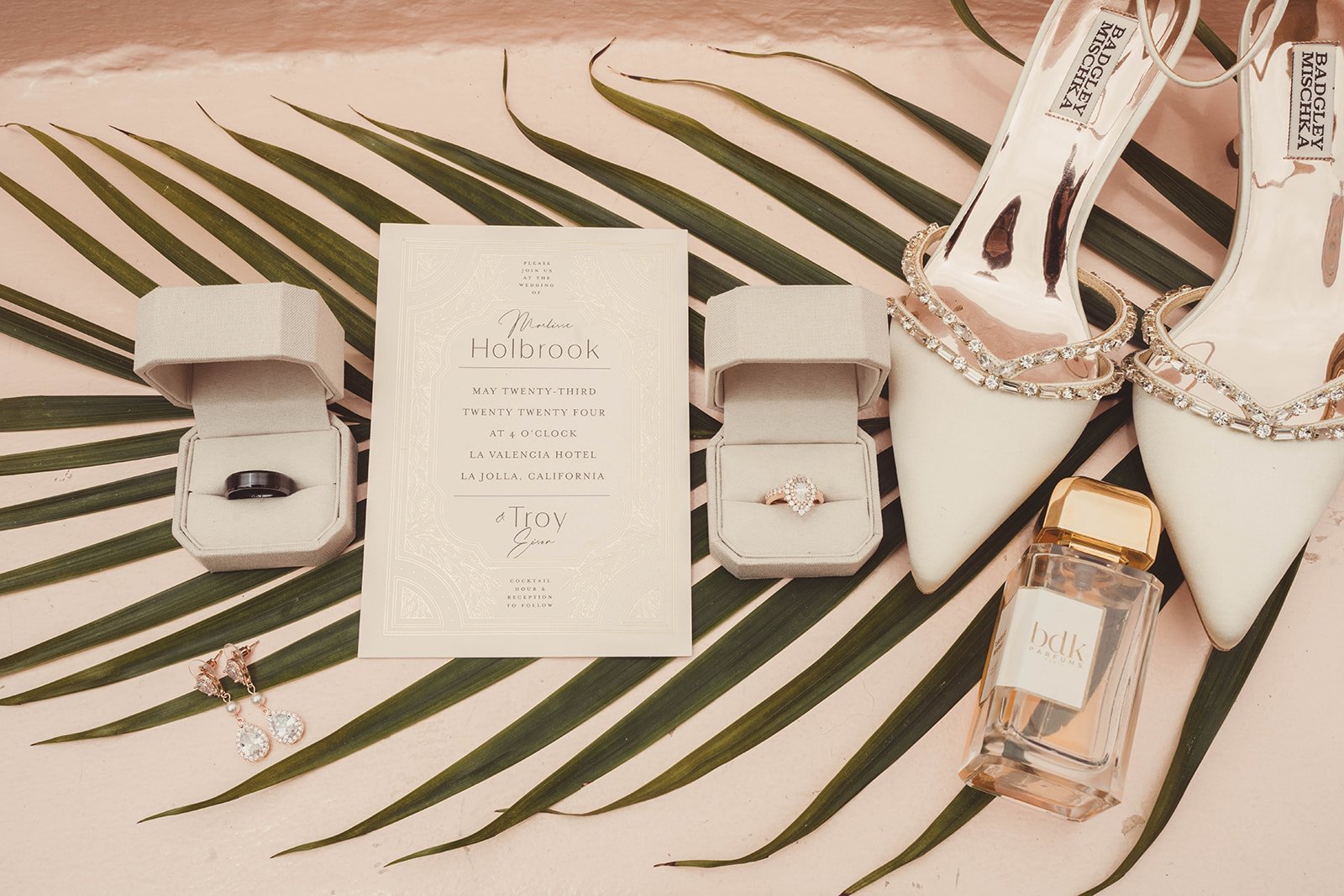 Wedding invitation flat lay with rings, earrings, shoes, and perfume on palm leaves and pink background.