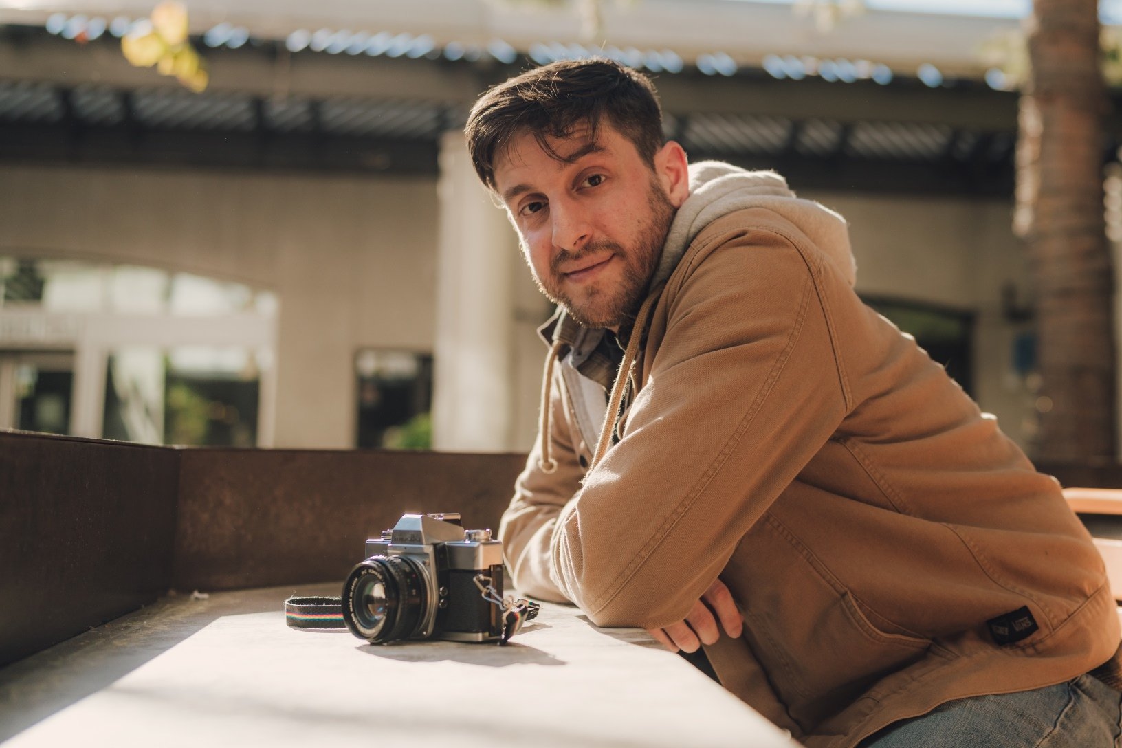 Man in a tan jacket leaning on a table outdoors with a vintage camera next to him.