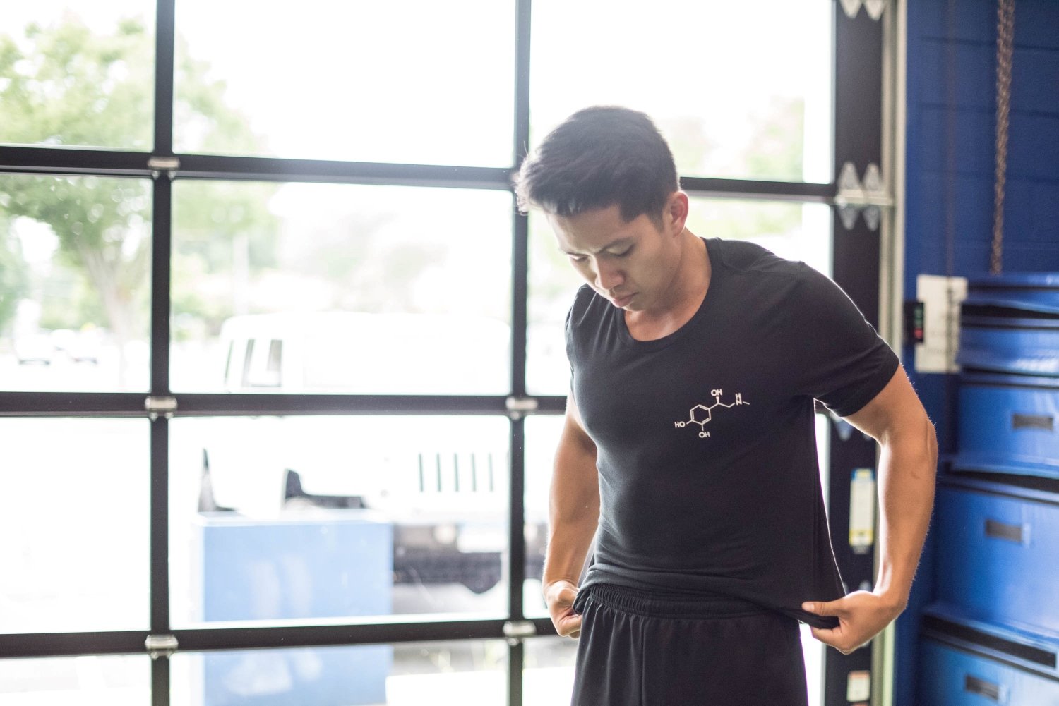 Man in gym holding black T-shirt with chemical structure design, garage door in background.