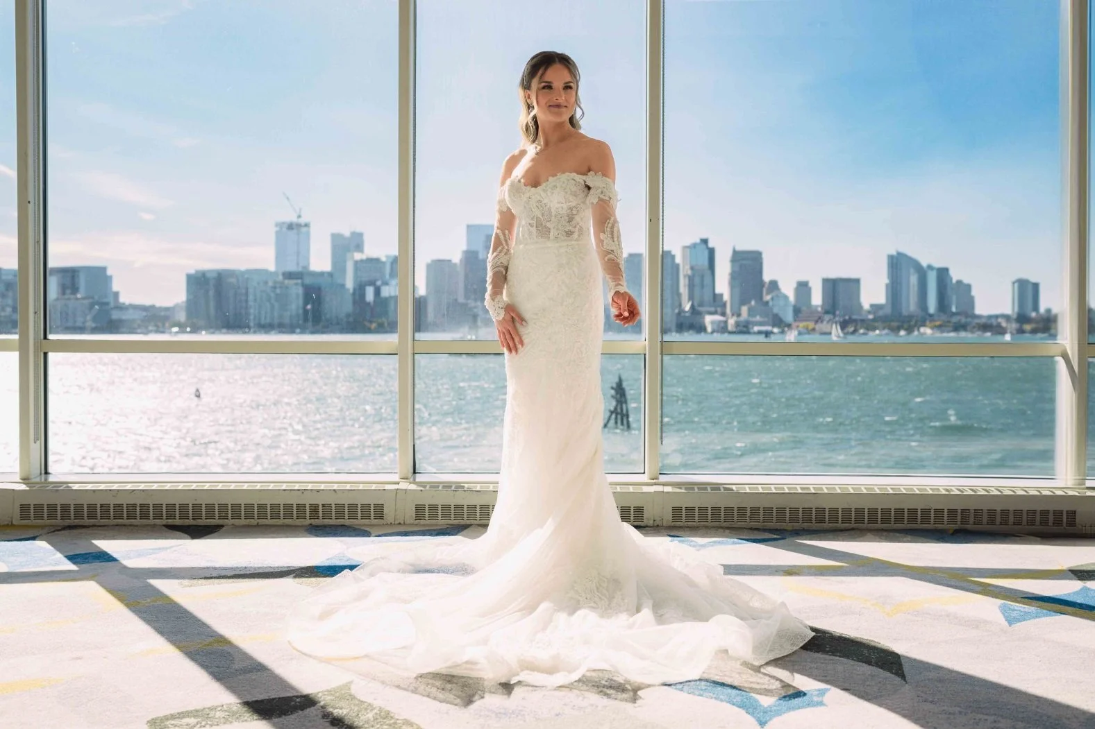 Bride in off-the-shoulder lace wedding dress stands indoors with city skyline and waterfront view.
