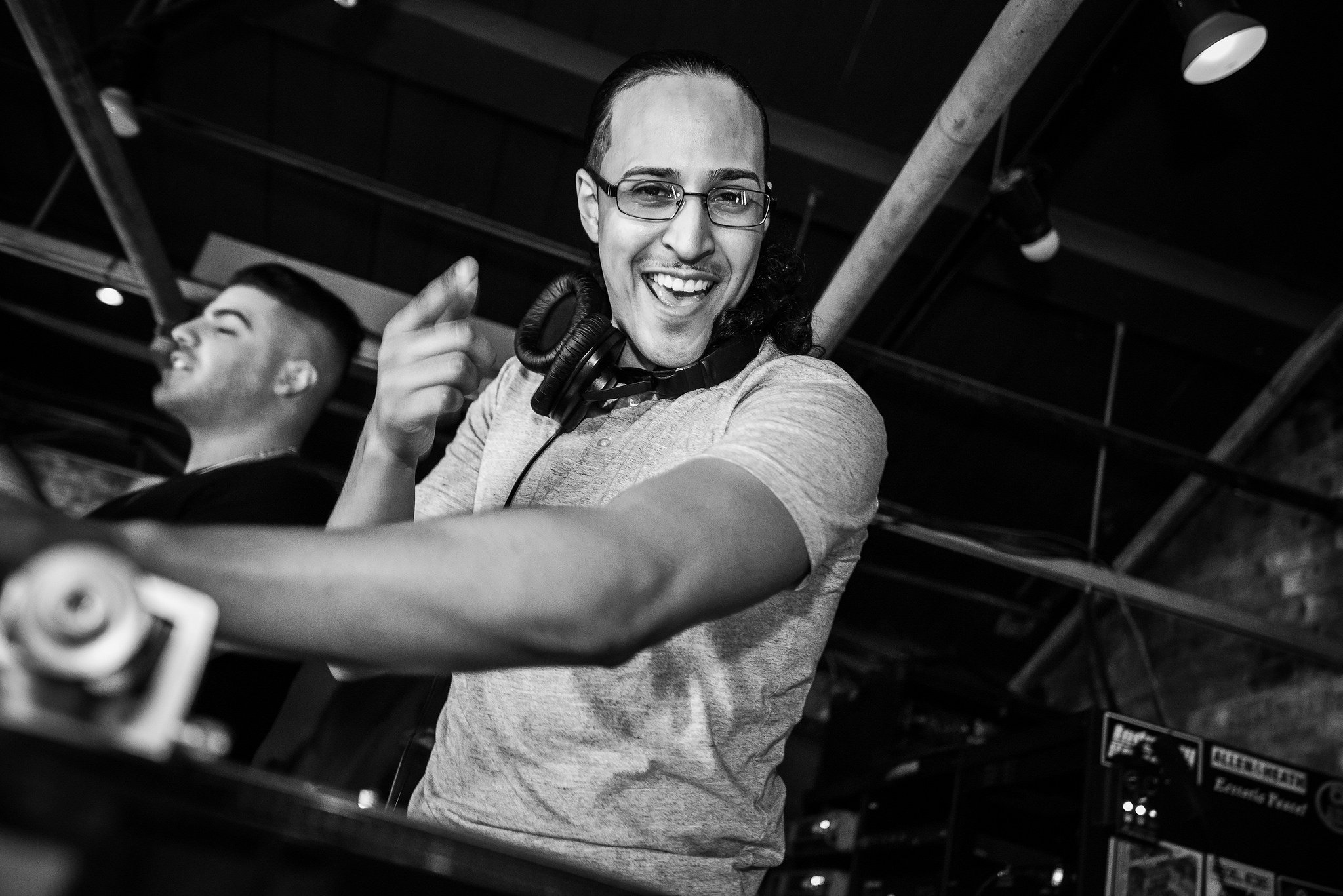DJ wearing glasses and headphones performing in a lively venue, smiling and gesturing energetically with another person in the background.
