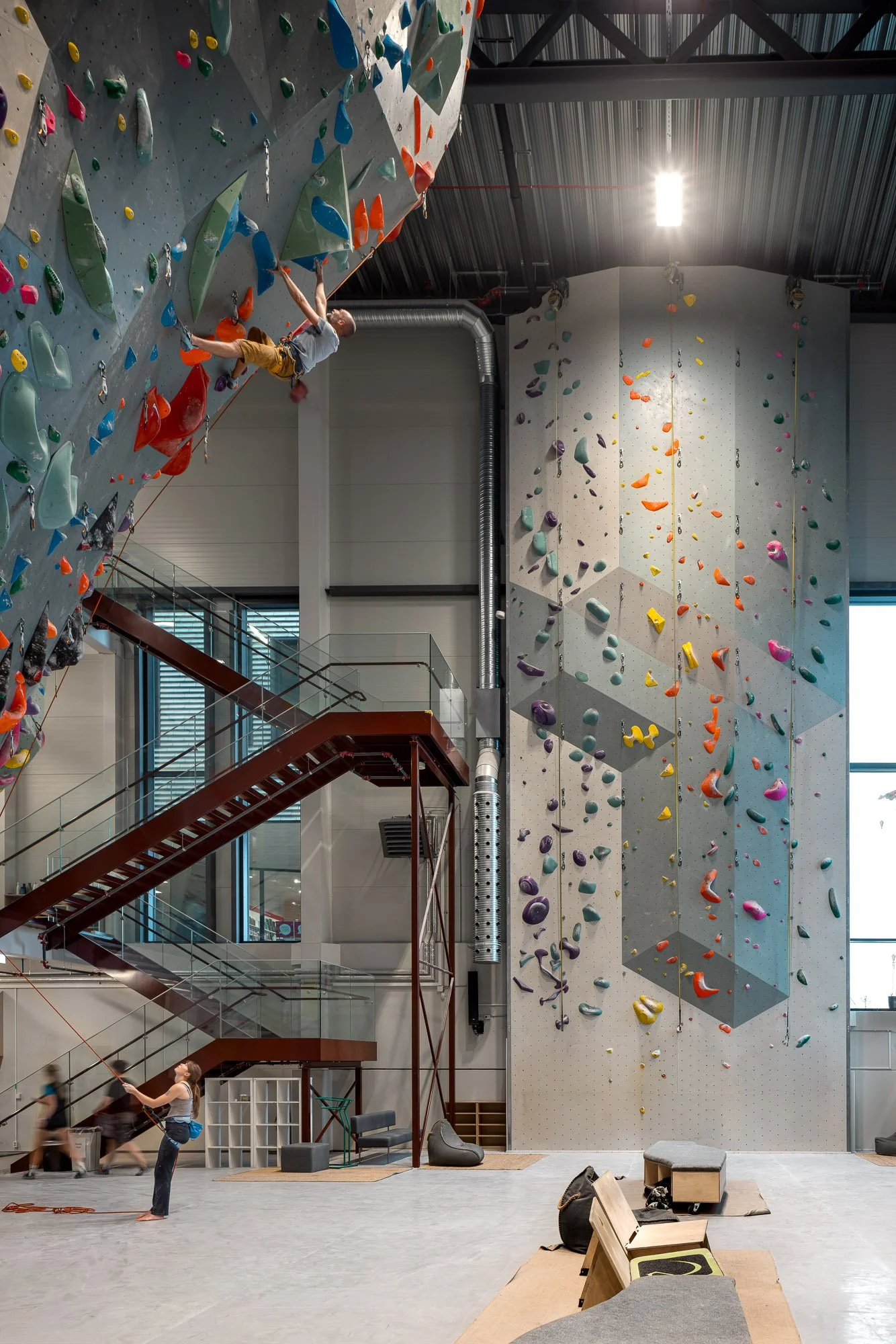 Innendørs klatrevegg med fargerike hånd- og fotgrep, en person som klatrer og en person som holder en sikkerhetsline, trapper og sitteplasser