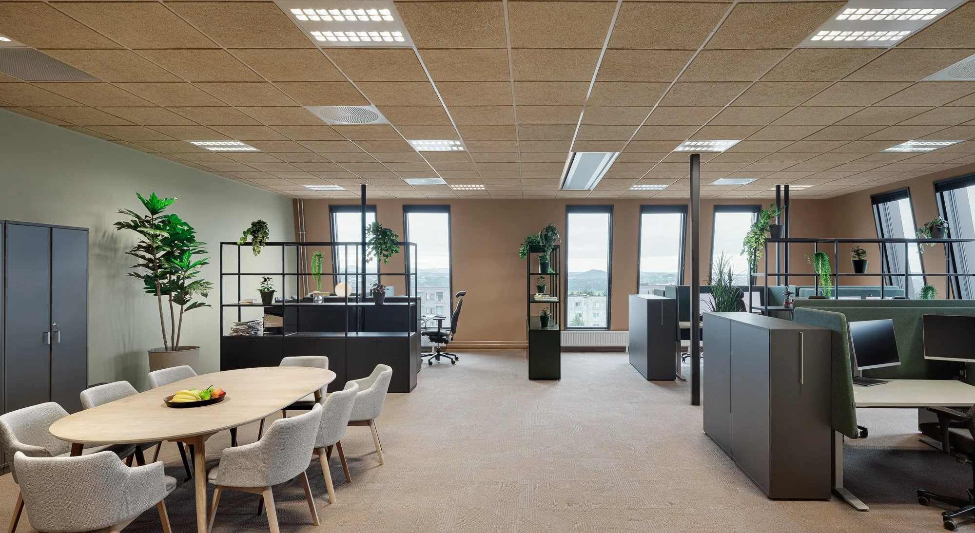 Et lyst og romslig kontorlokale med store vinduer, flere kontorplasser med datamaskiner, grønne planter og et møtebord med stoler