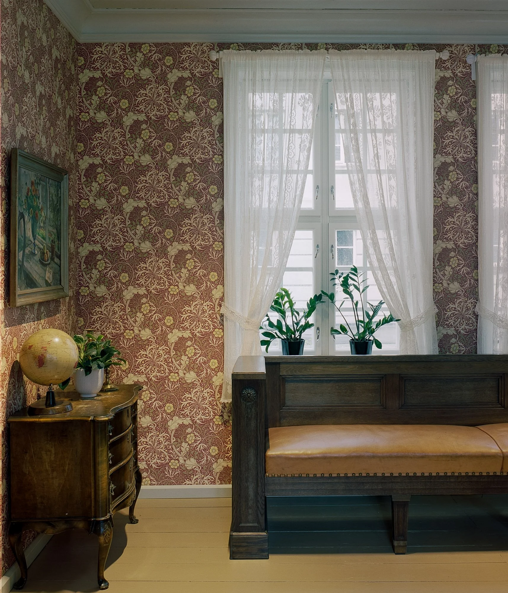 Et rom med tresnekke, to planter på vinduskarmen, lyst gardin, og tapet med blomster- og bladmønster.