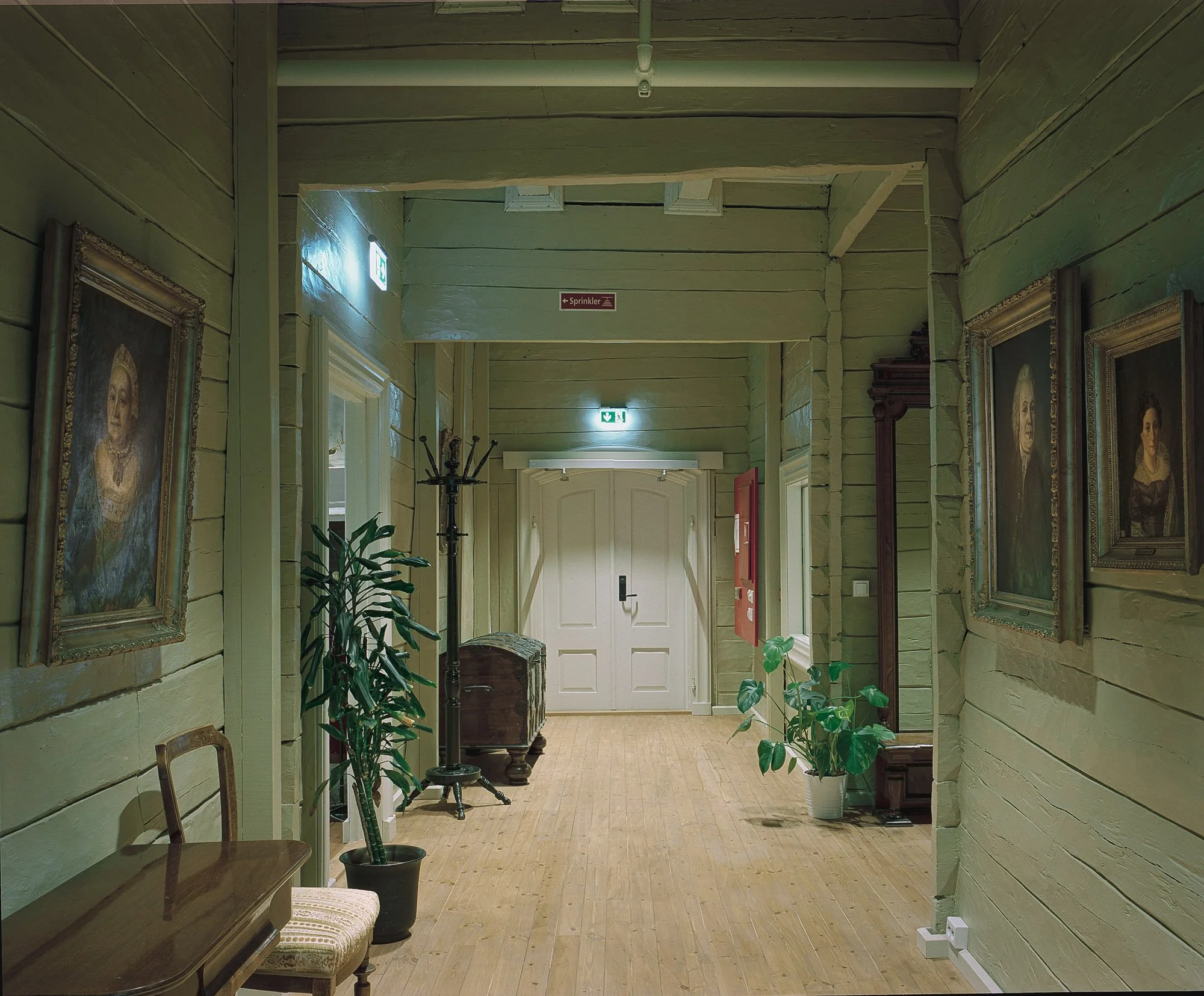 Innendørs gang med grønne vegger, treverk, kunstmalerier, planter, og møbler.