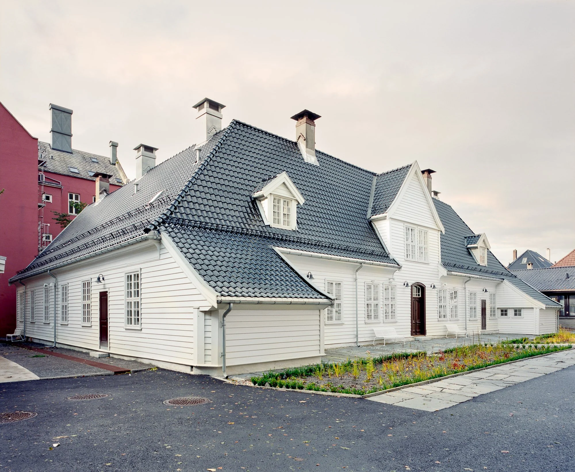 Hvitt trehus med svart tak, flere små takvinduer og hvite vinduer, omgitt av en liten hage med blomster og en parkeringsplass foran.