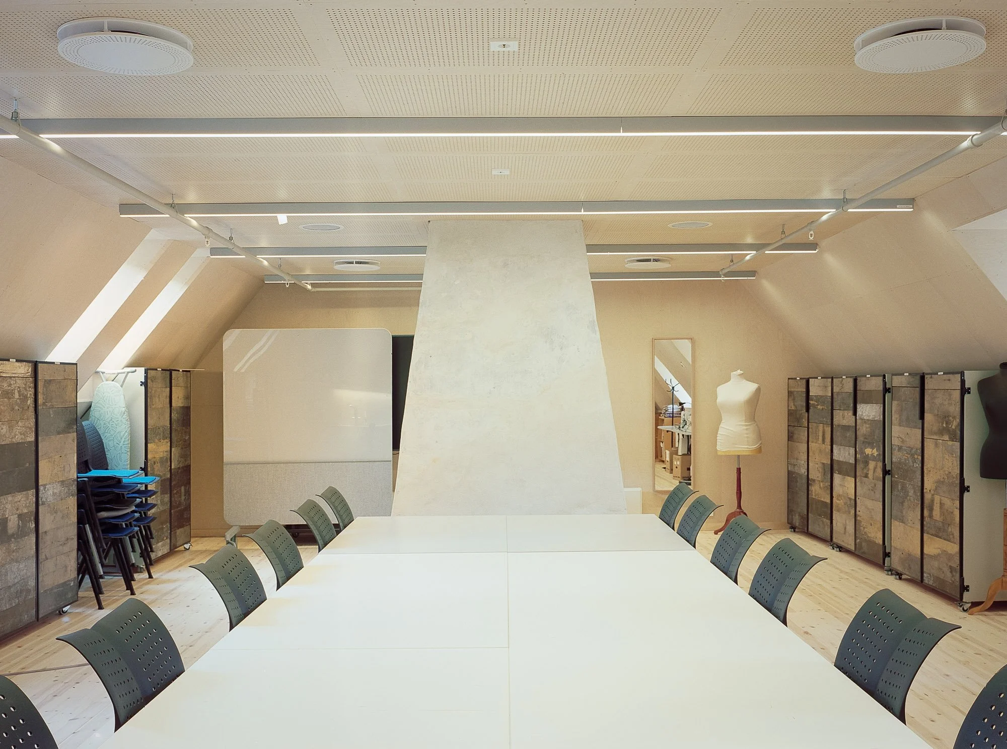 Innendørs møte- eller konferanserom med en lang hvit bordflate og flere grønne stoler rundt. På venstre side er det stablede stoler og møbler. På høyre side er det utstillinger av tekstiler og mannekster. Taket har hvite runde lyspærer og synlige ven