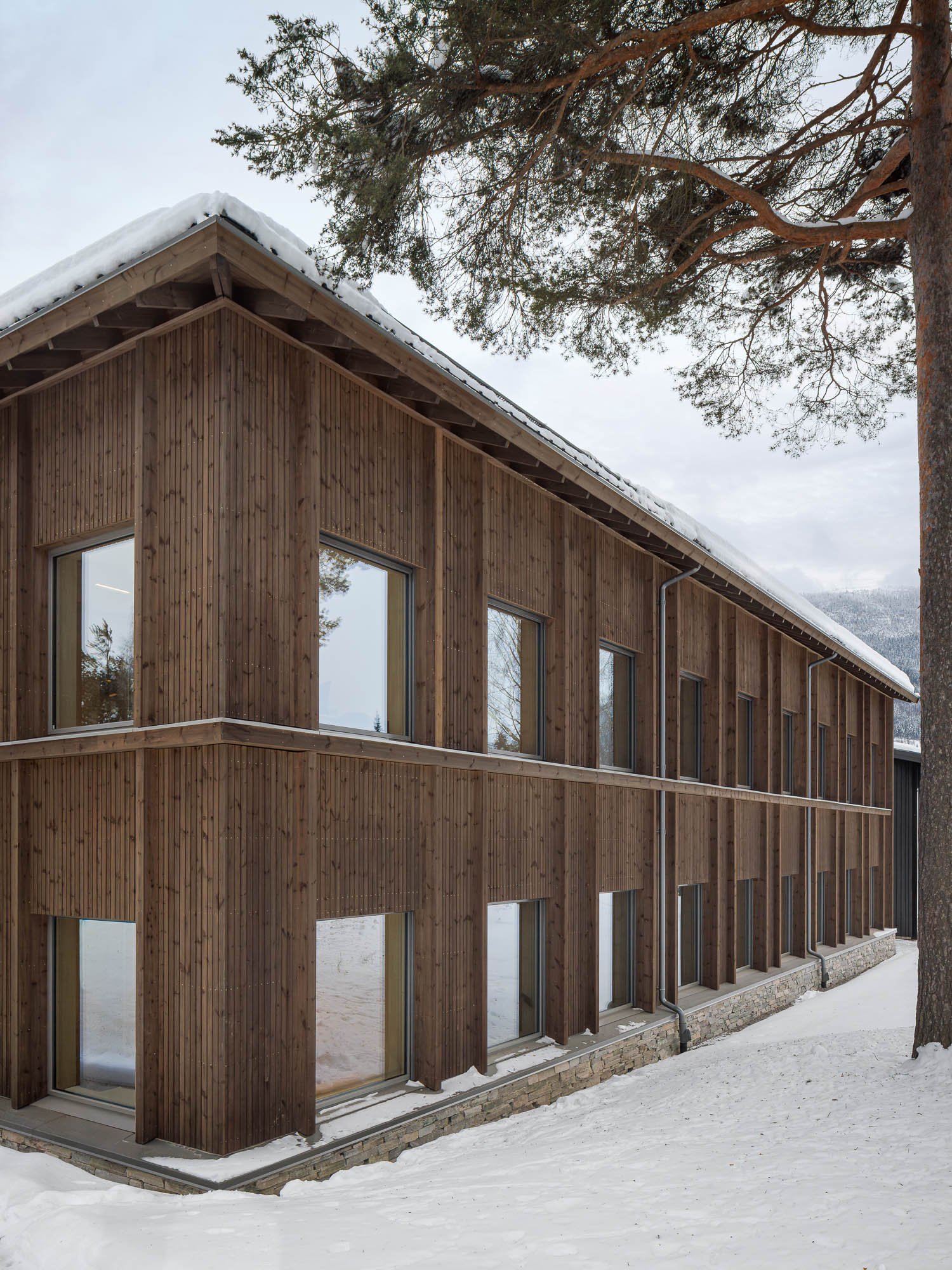 Et trehus med tre etasjer, kledd i trepanel, omgitt av snø, sett fra utsiden, med store vinduer og et tre i forgrunnen.
