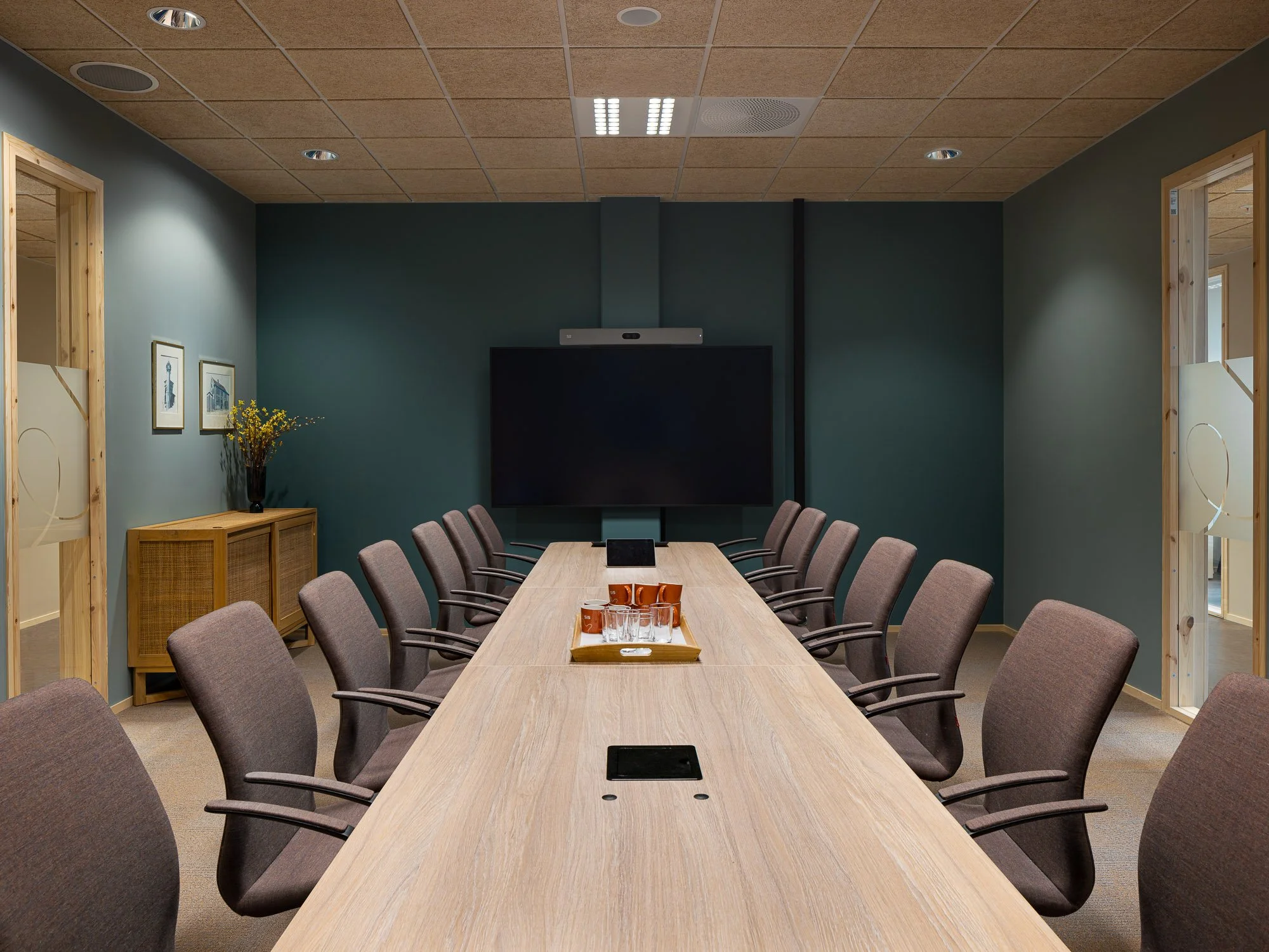 Et konferanserom med lang ovalt bord omringet av stoler, en stor skjerm på veggen og et videokonferansesystem over skjermen.