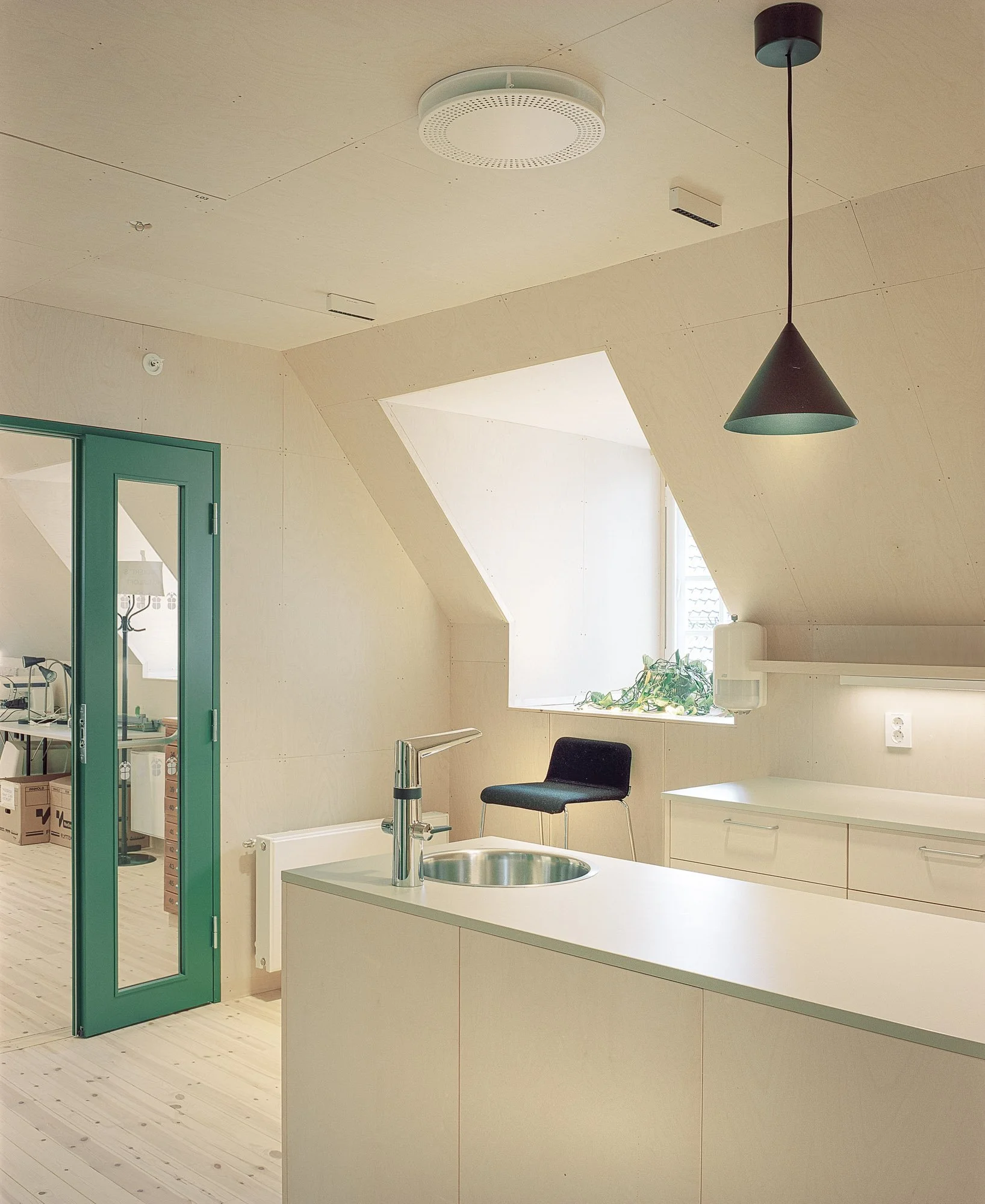 Moderne kjøkken med hvit benkeplate, vask, svart stol, skjult vindu, grønne dørkarm, trepanelvegger, svart taklampe og takvindu.