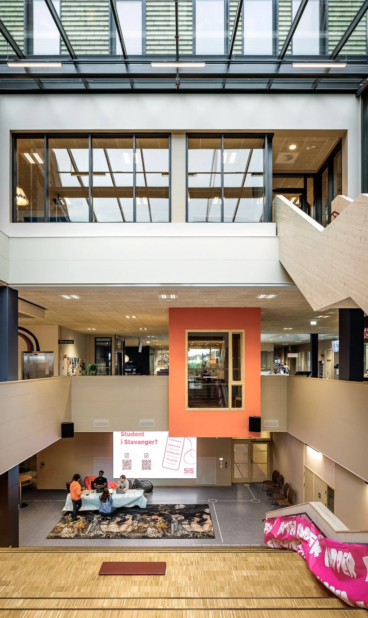 Innendørs plass med et lavere og et øvre etasjenivå, slik som et bibliotek eller kontorlokale, med flere mennesker, et bord med tre personer og en plakat med teksten 'Student i Stavanger?'.