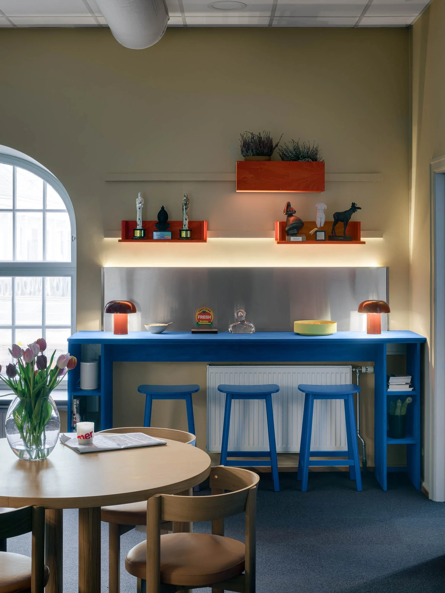 Interiør av et rom med en blå bardisk, tre blå stoler, en rund trebord med blomster i en vase, og hyller med diverse objekter og dekorasjoner.