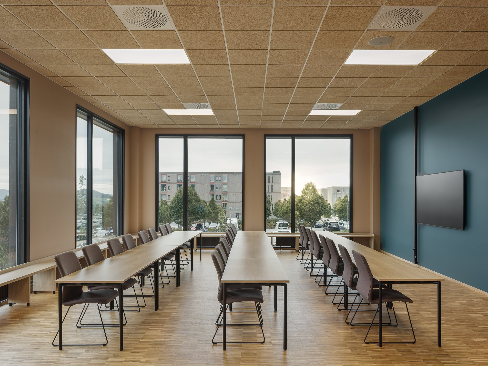 Et møterom med lange trebord og stoler, store vinduer og en vegg med TV-skjerm, med utsikt over utendørs parkeringsområde og bohus.