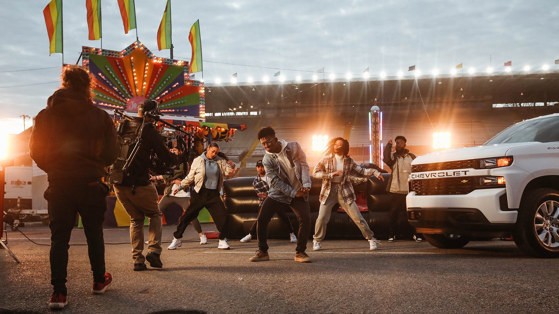 Group of dancers performing on a racetrack at sunset, with carnival rides and flags in the background, and film crew capturing the scene.