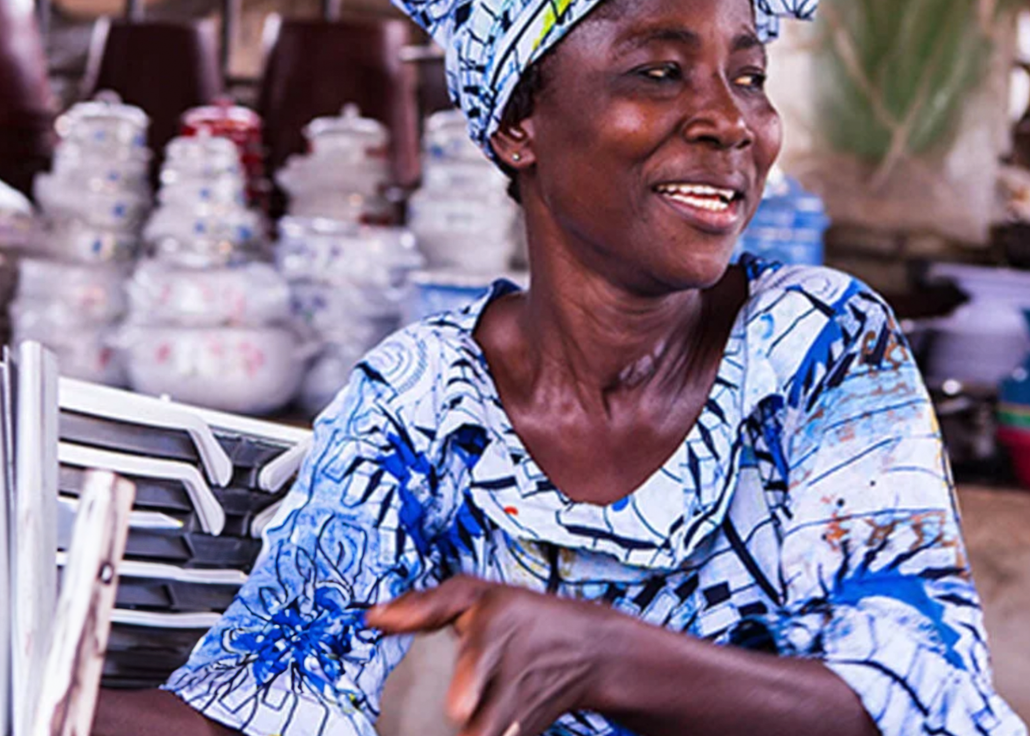 A smiling woman in a blue and white patterned dress and headwrap sitting at an outdoor market or shop, with stacks of bowls and other items in the background.
