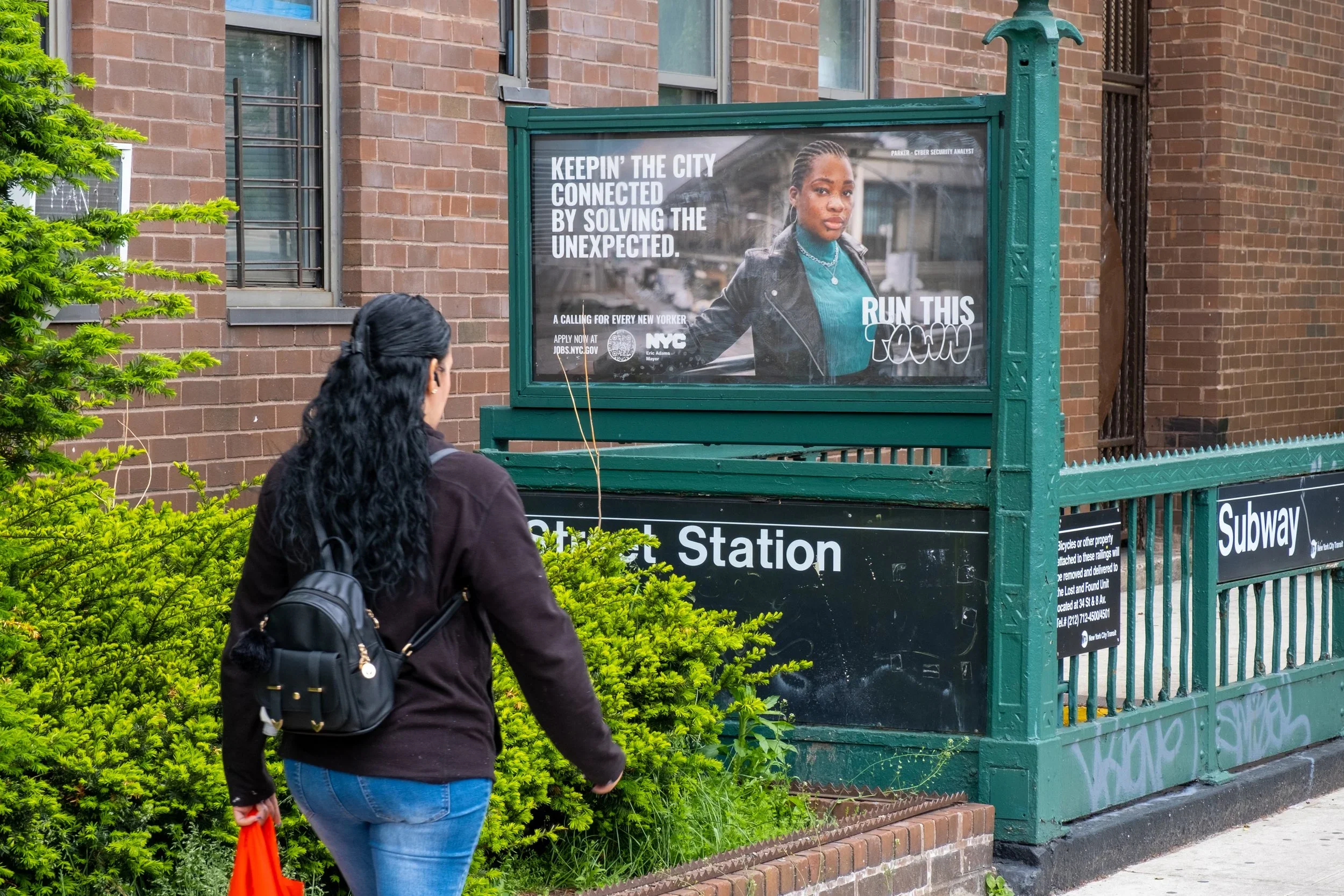 A woman with black hair, wearing a dark jacket and blue jeans, holding an orange bag, standing near a green subway entrance and looking at an advertisement on a city subway station sign.
