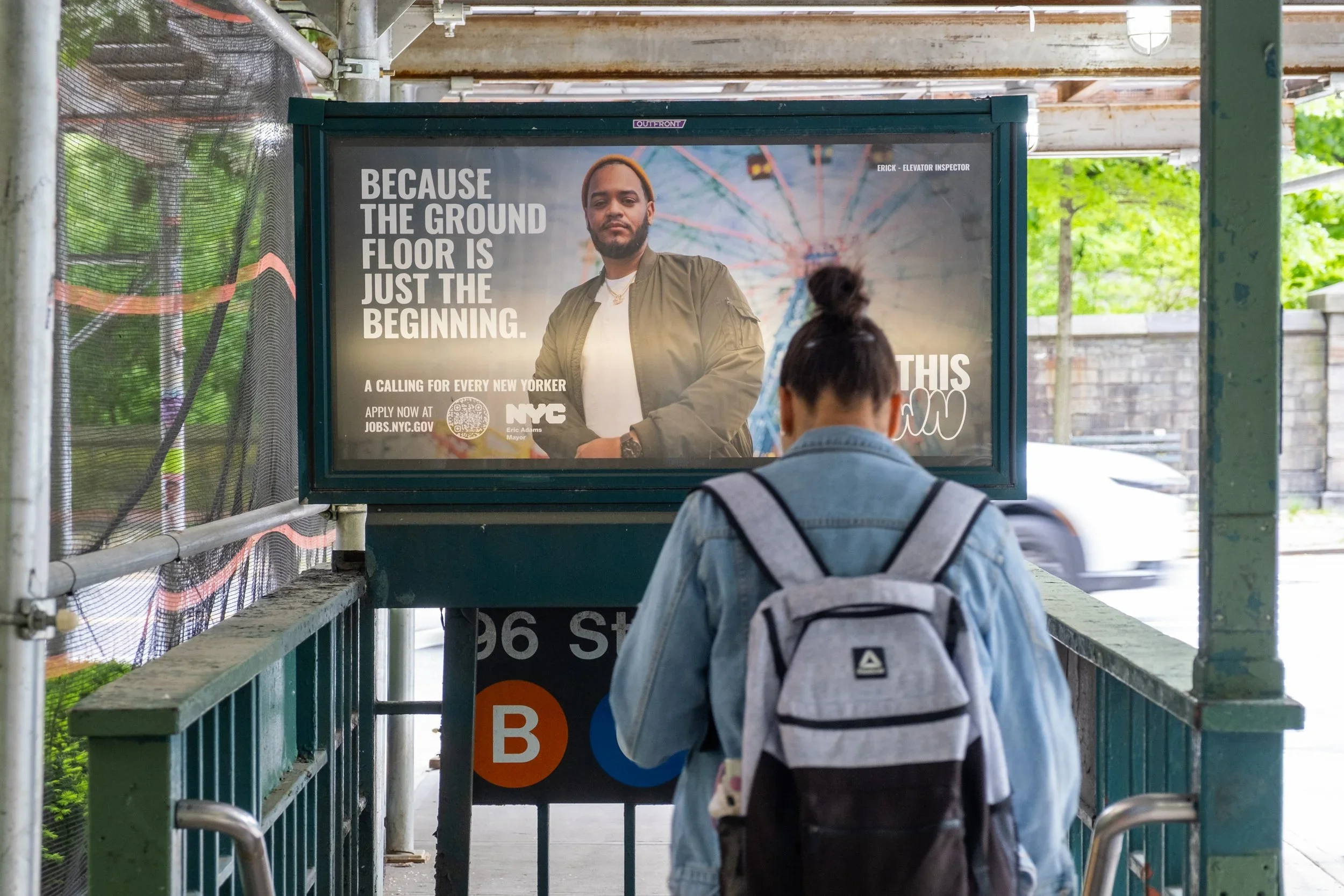 A person with a gray backpack and denim jacket is standing at the entrance of a subway station, looking at an advertisement poster. The poster features a man with a beard wearing a green jacket, with the text 'Because the ground floor is just the beginning' and NYC branding. There are subway signs for B train underneath.