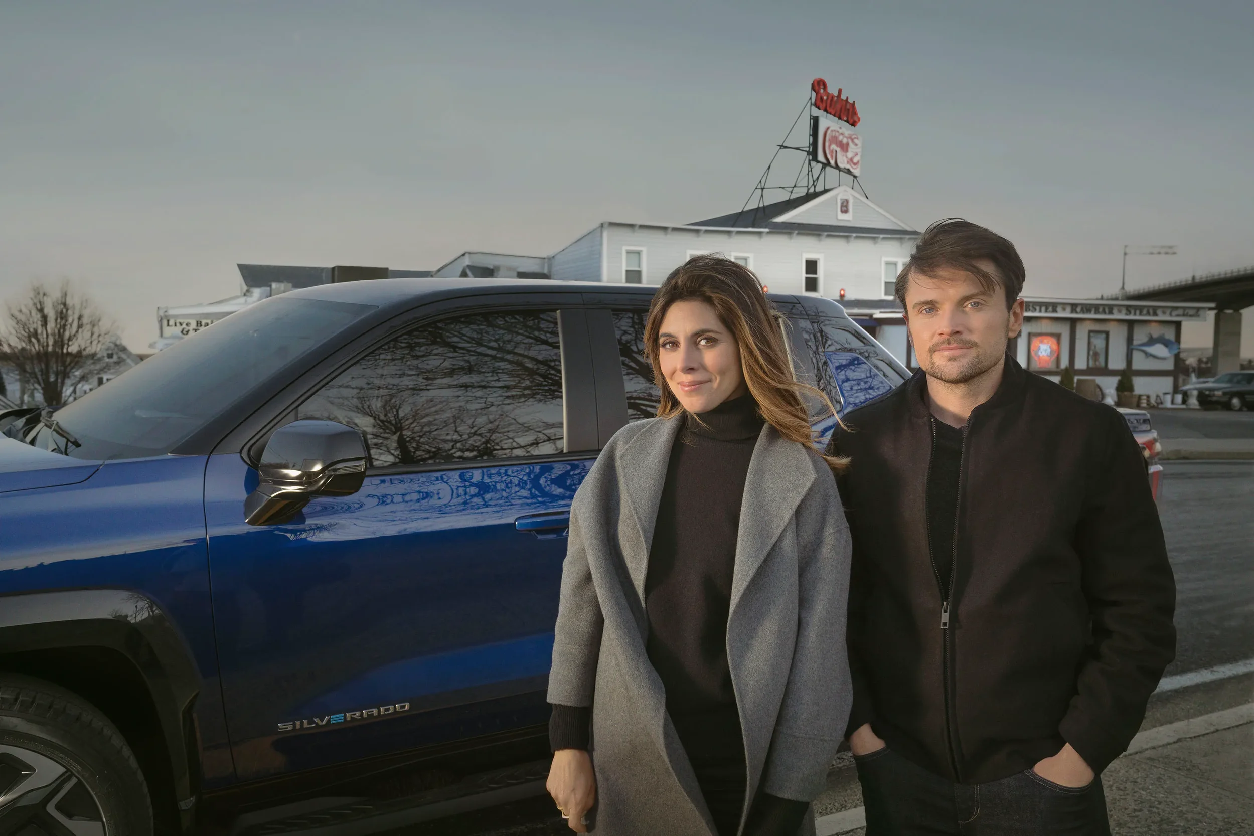 A woman and a man standing next to a blue Chevrolet Silverado pickup truck in an outdoor parking lot. The woman is wearing a gray coat and black top, and the man is wearing a black jacket. In the background, there is a building with a large red sign and some trees.