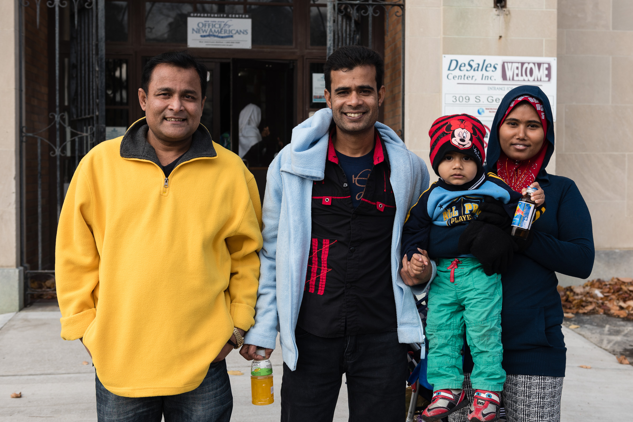   Nor Kalimullah (center) stands with his friend Abdul Alim (left) and Alim’s wife and child, Jaura and Abdul (right), outside the entrance to the Mohawk Valley Resource Center for Refugees in Utica, New York.&nbsp; Joseph Ryder   