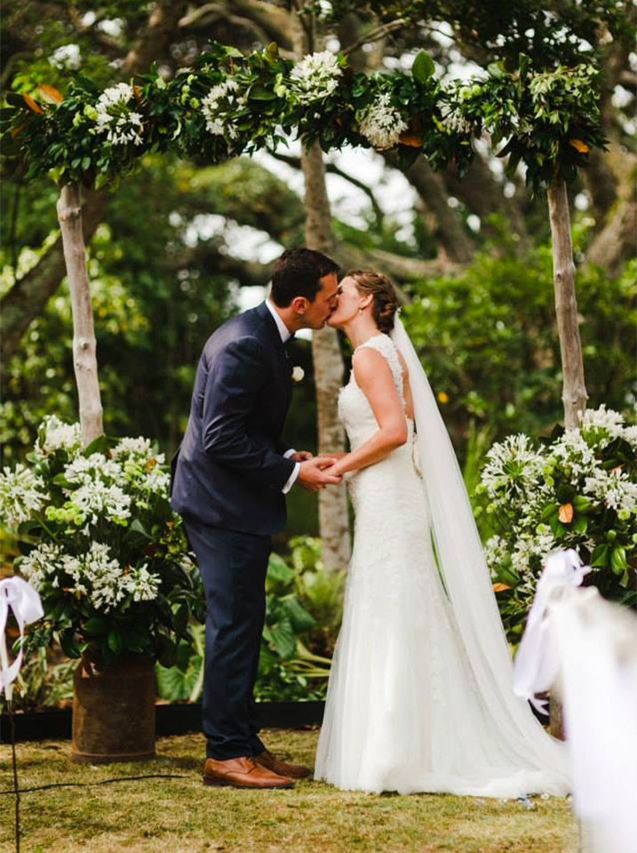 Couple kiss under arch decorated with flowers by Wildflowers Coromandel