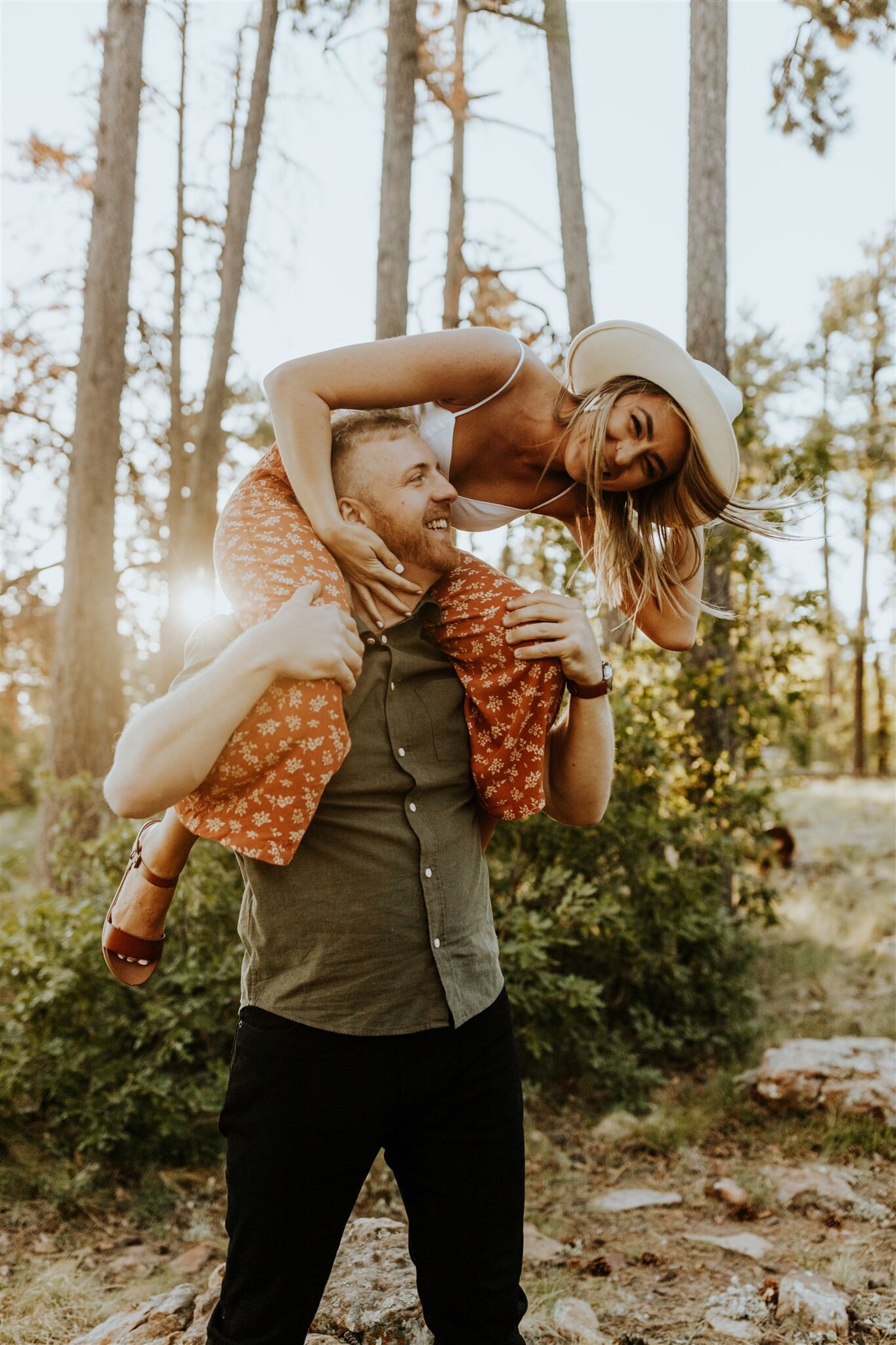  playful photo of a woman in a hat on a man’s shoulders, they are smiling at one another 