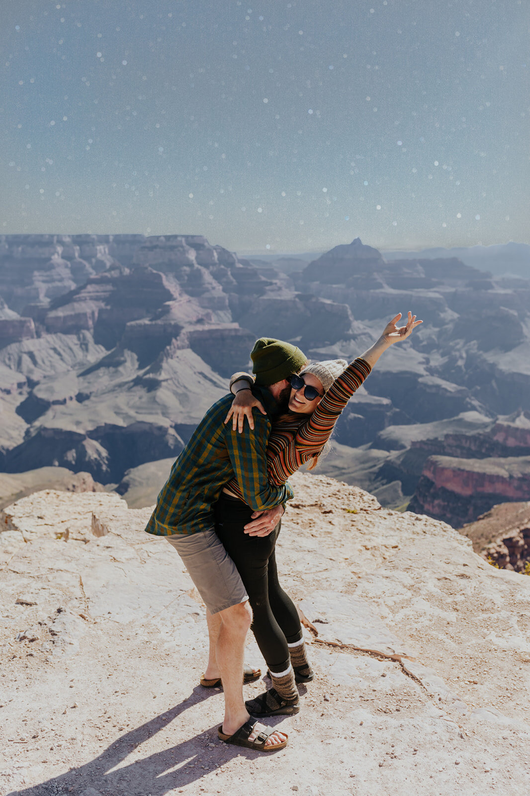  a woman stands triumphantly and smiling on a cliff while a man kisses her cheek 