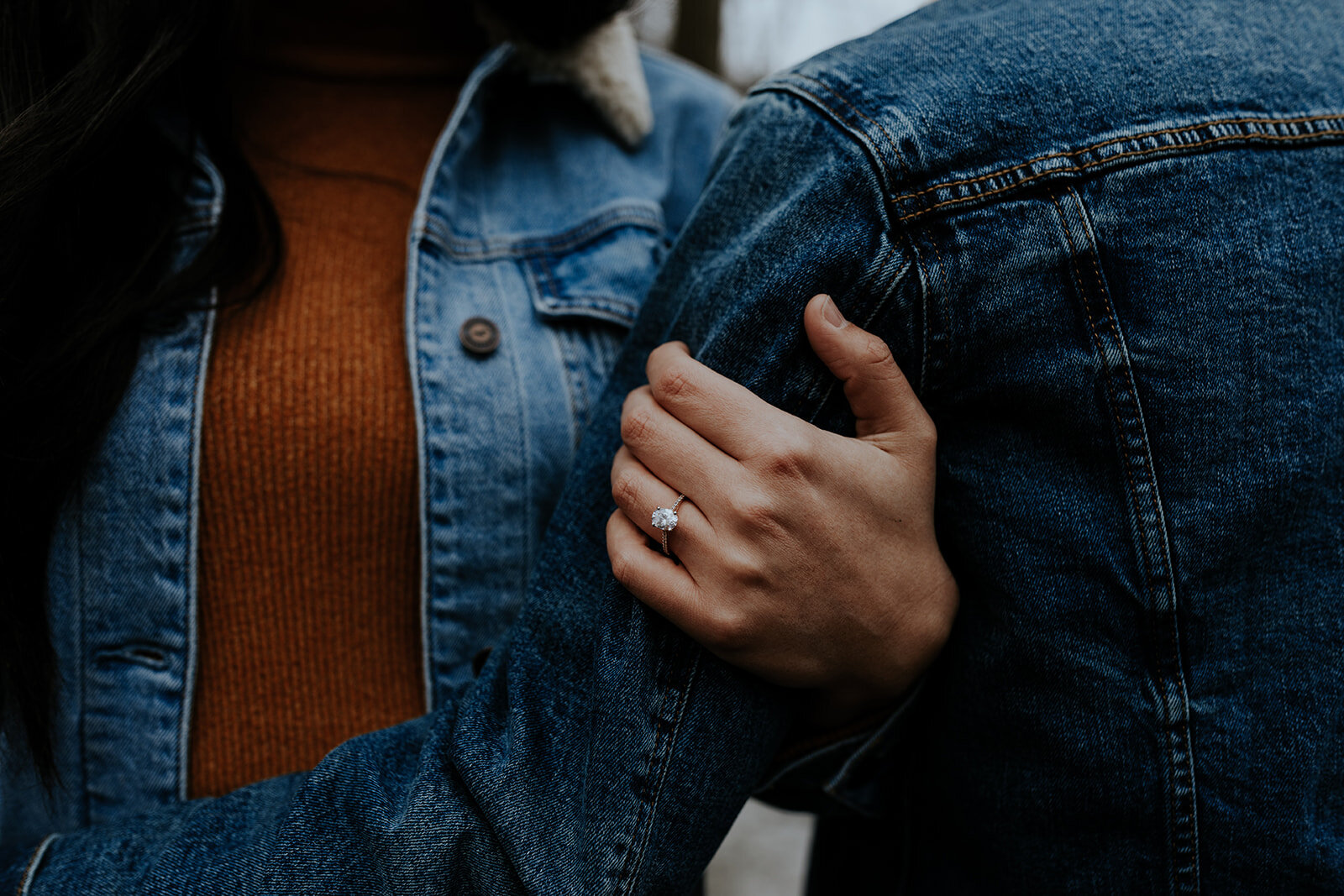  close up shot of couple in jean jackets and a women’s hand with an engagement ring grasping her lover’s arm  