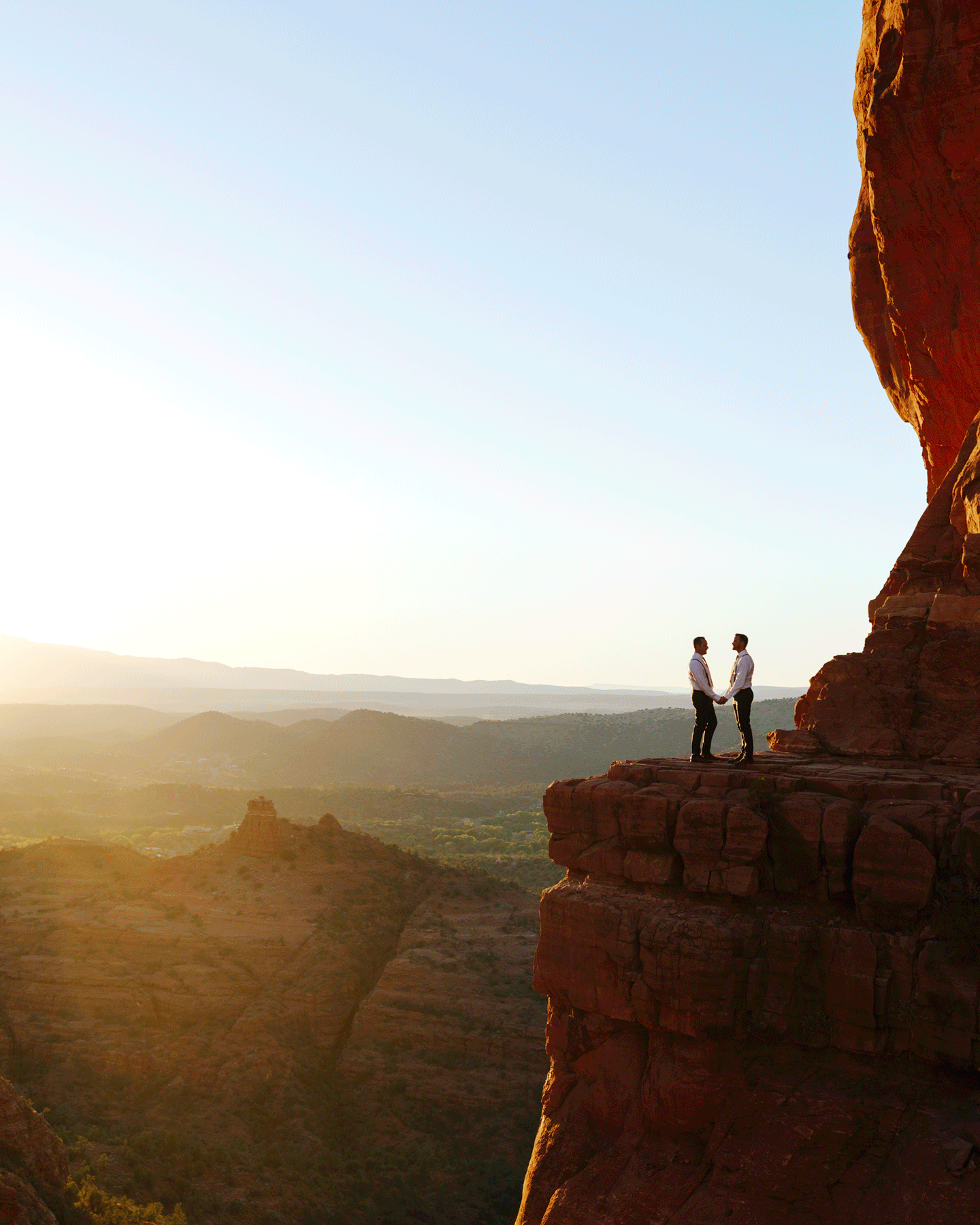 Two people holding hands on a cliff with a scenic desert landscape at sunset.