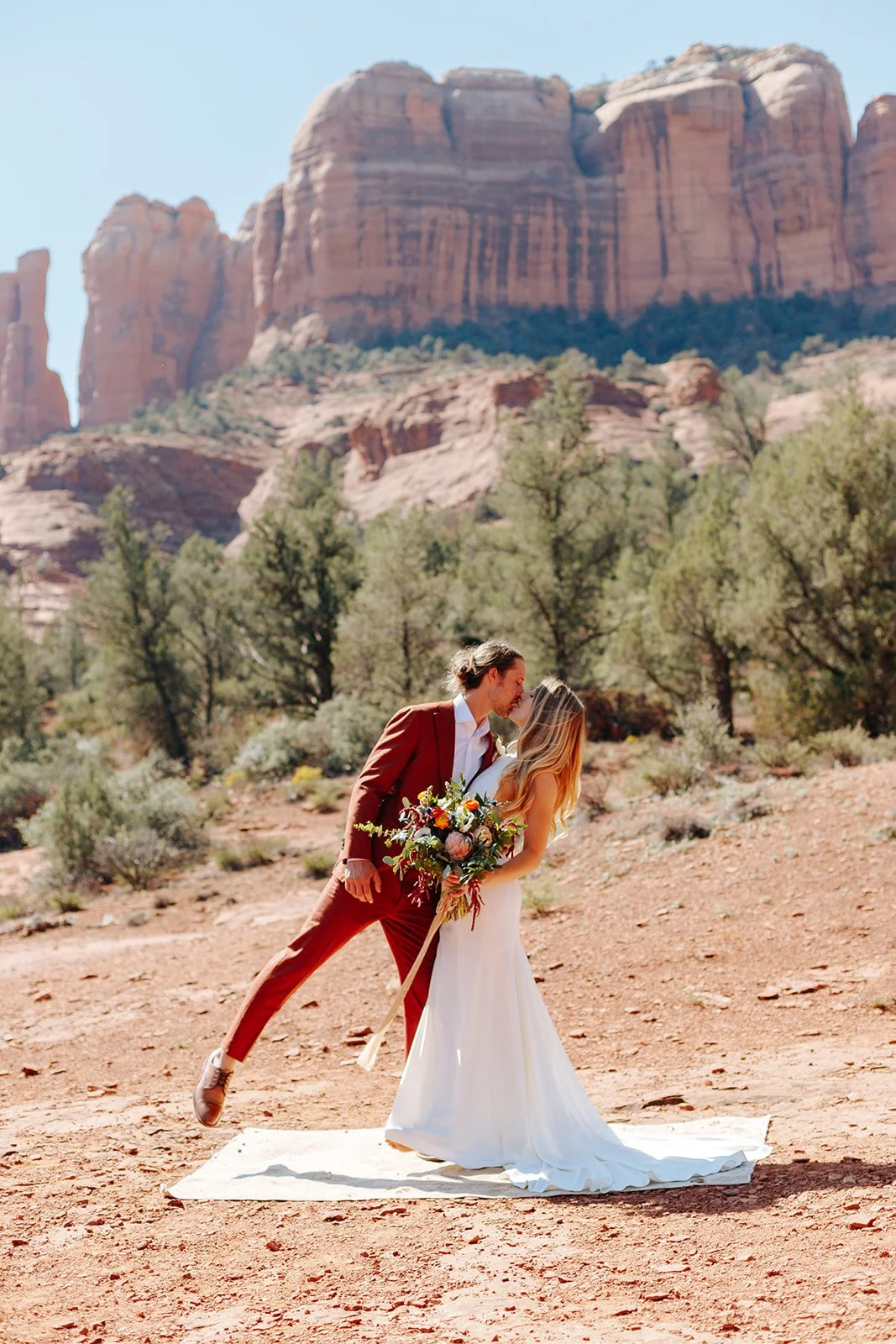 Couple in wedding attire kissing in desert landscape with red rock formations.