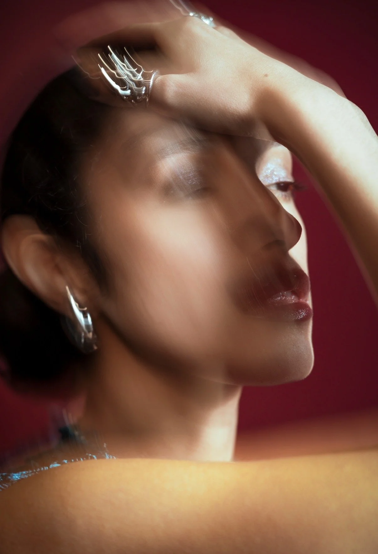 Layered side-profile portrait of Melody V with one hand above her forehead, wearing silver earrings against a red background.