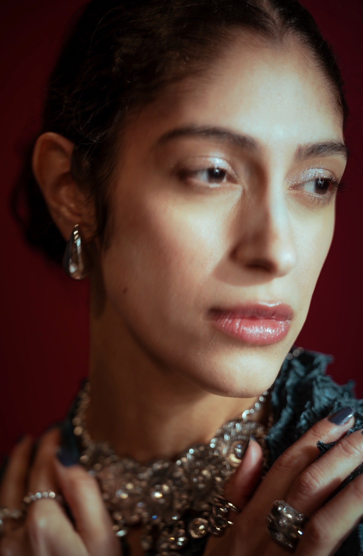 Close-up portrait of Melody V against a deep red background, wearing silver jewelry with her hands near her shoulders.