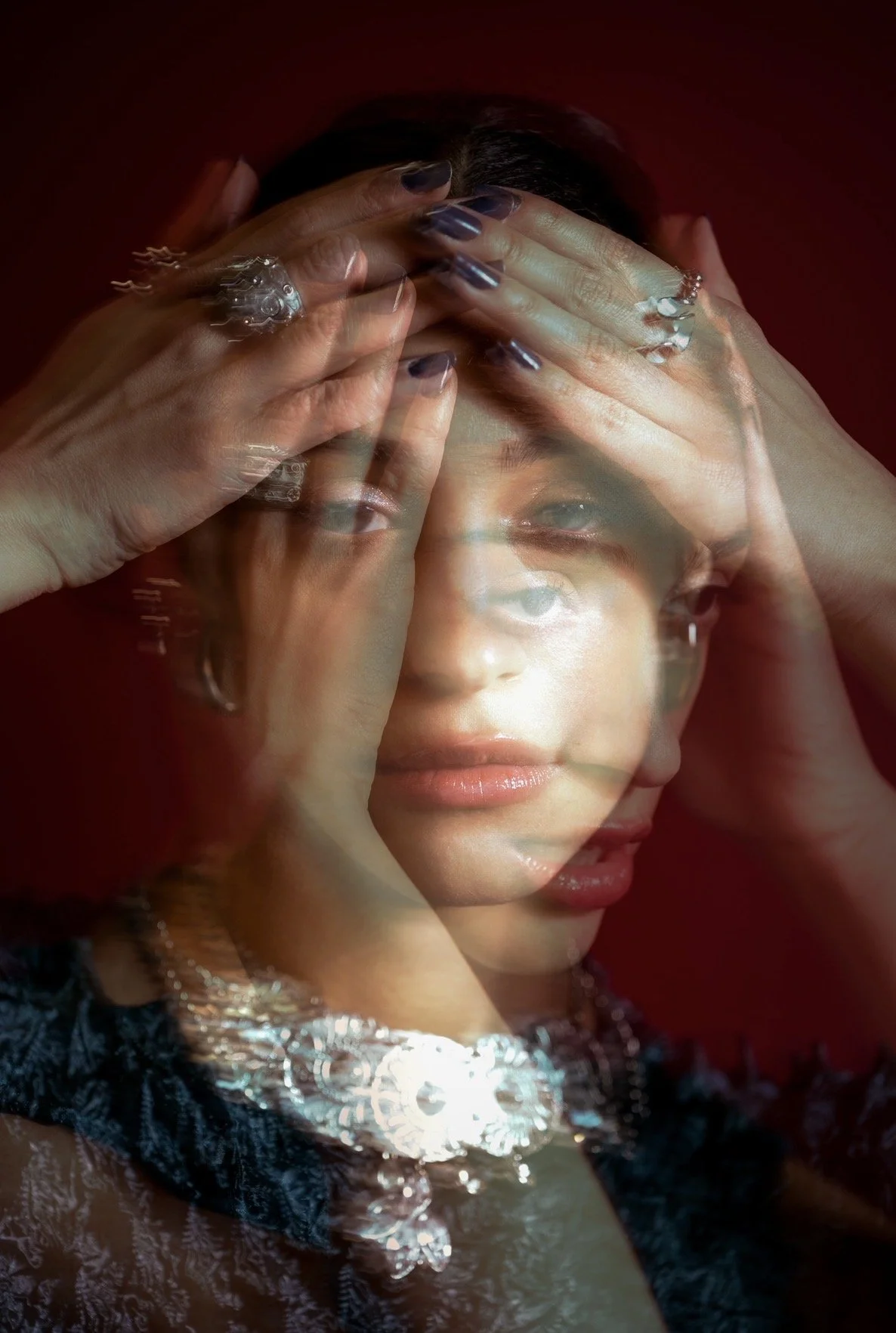 Layered exposure portrait of Melody V with her hands across her forehead and silver jewelry against a dark red background.