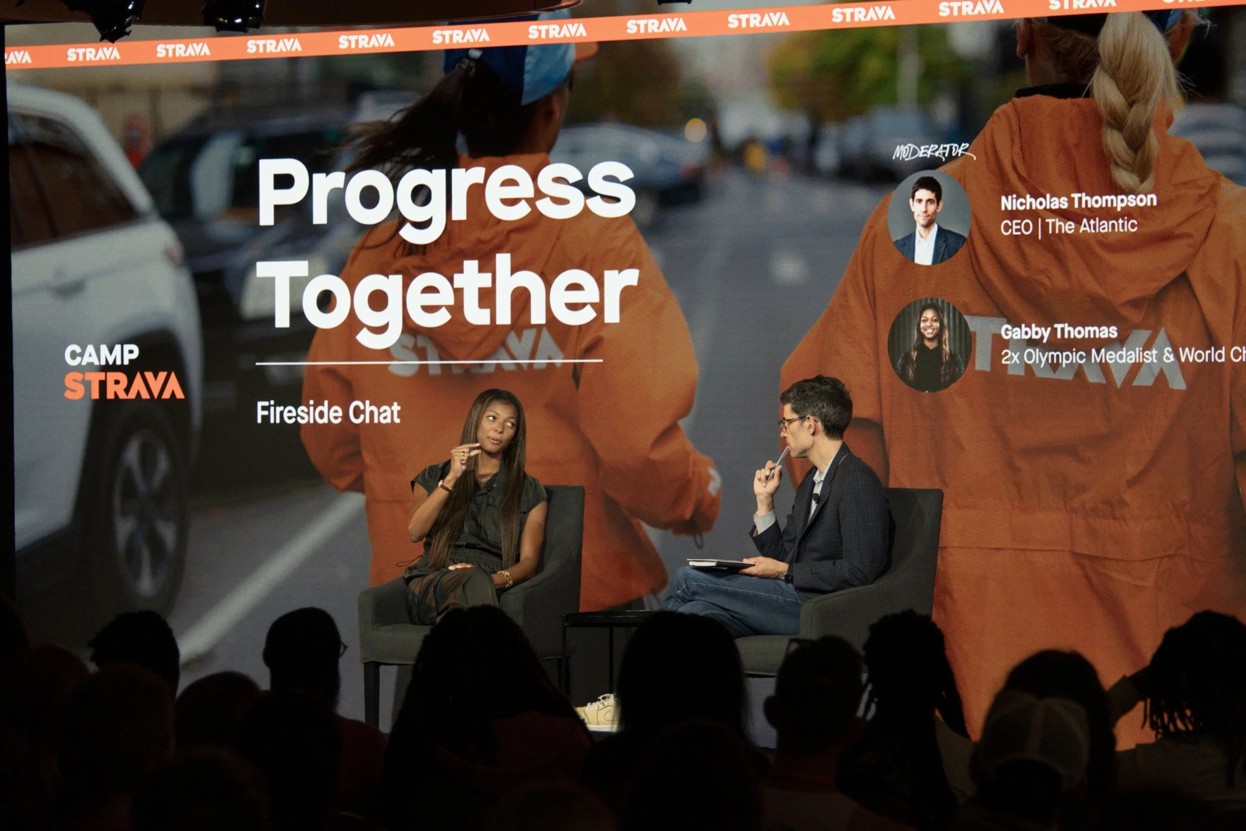 A panel discussion at a conference with two people seated on stage and a large screen behind them displaying the words "Progress Together" and images of speakers Nicholas Thompson and Gabby Thomas, with the Strava logo and event title "Camp Strava".