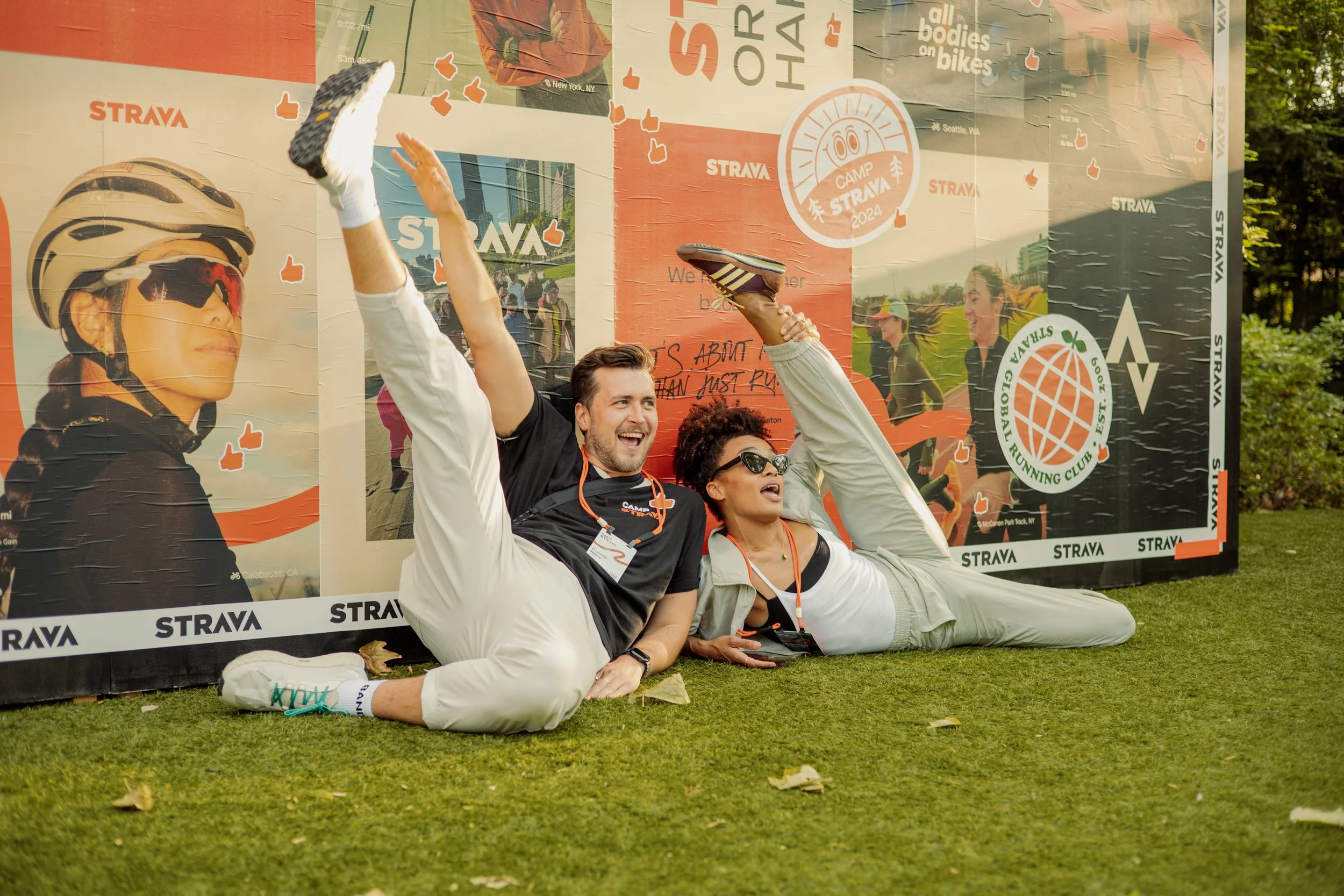 Two people are relaxing on the grass at a cycling event, with a large promotional backdrop behind them. The background features various logos and images related to cycling and running, including event branding for Strava and Camp Strava 2024.
