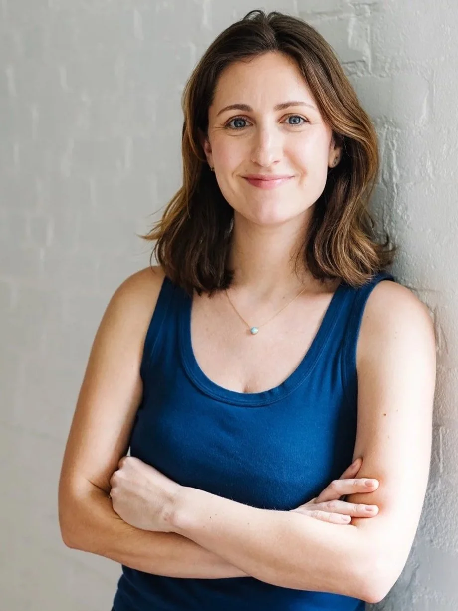 A woman with shoulder-length brown hair and blue eyes, smiling softly, wearing a blue sleeveless top and a dainty necklace, standing against a white brick wall with arms crossed.