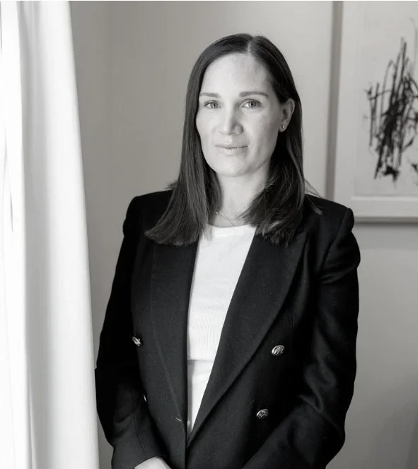 Black and white photo of a woman with shoulder-length dark hair, wearing a dark blazer over a light-colored top, standing indoors near a curtain and a framed abstract artwork.