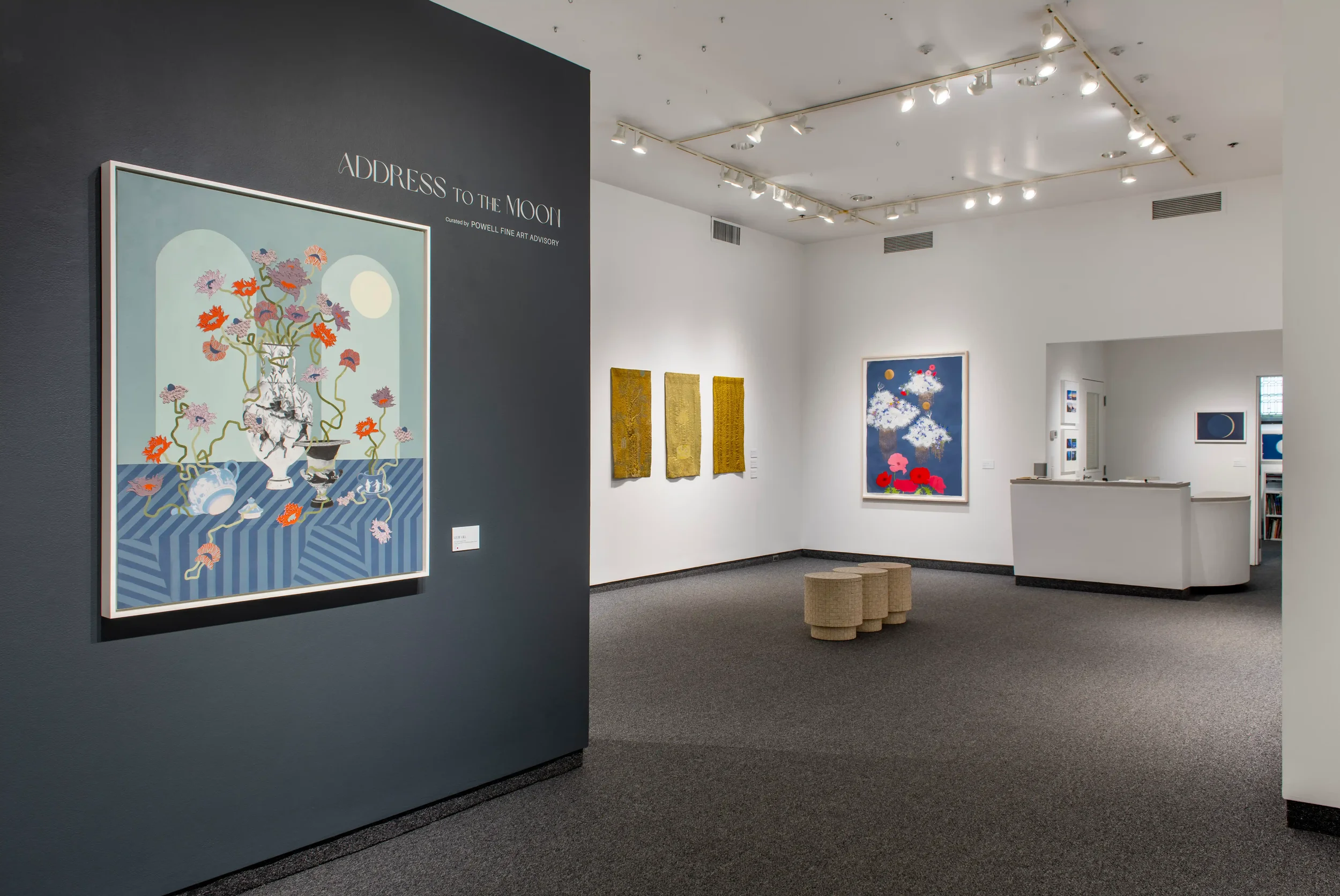 An art gallery showcasing various colorful paintings, including one titled 'Address to the Moon' on a dark wall, with additional unframed artworks on a white wall in a bright, modern space with ceiling lighting and a small seating area.