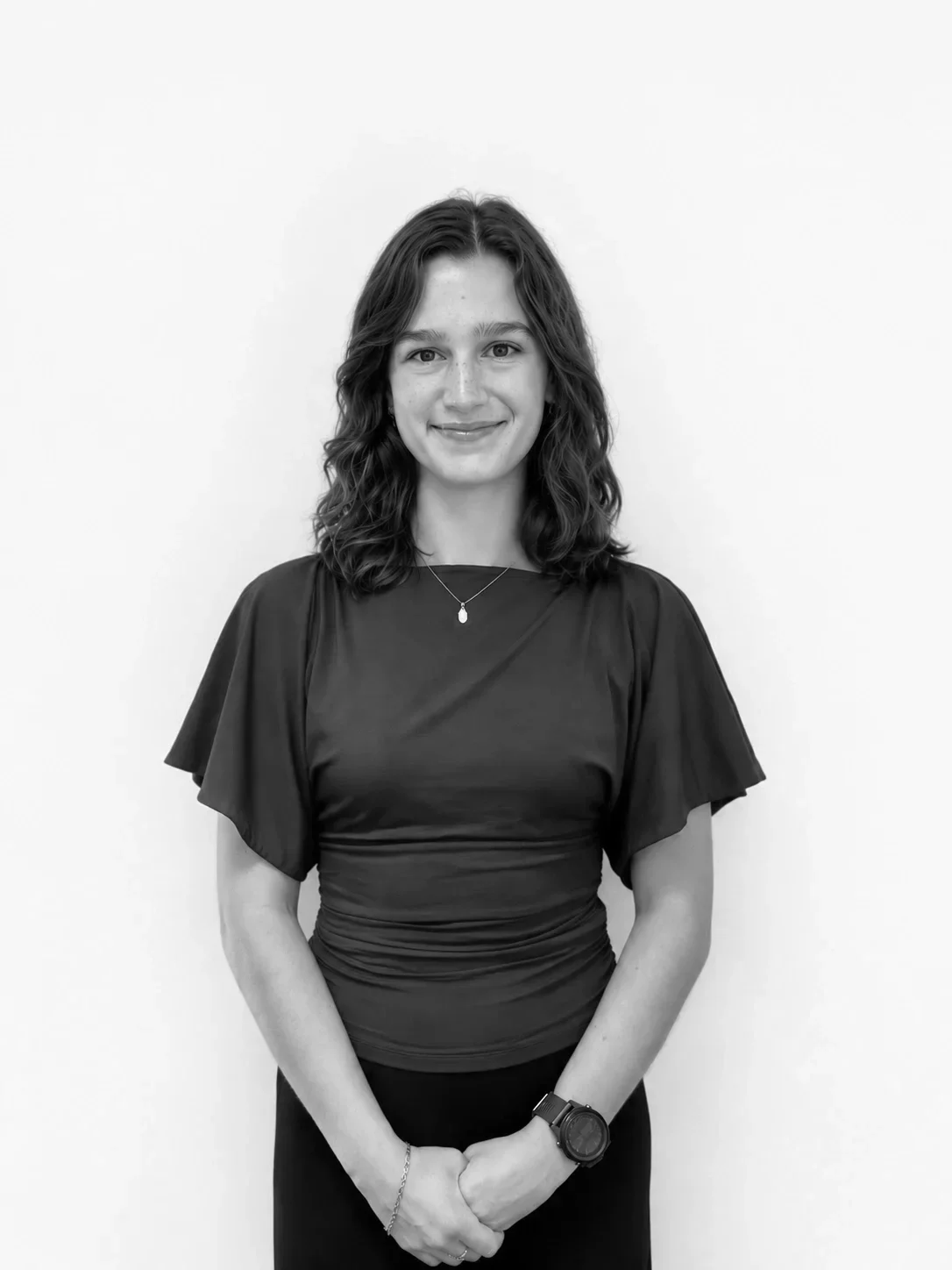 Black and white portrait of a young woman with wavy shoulder-length hair, smiling, wearing a short-sleeved top, a necklace, a watch, and a bracelet, standing against a plain white background.
