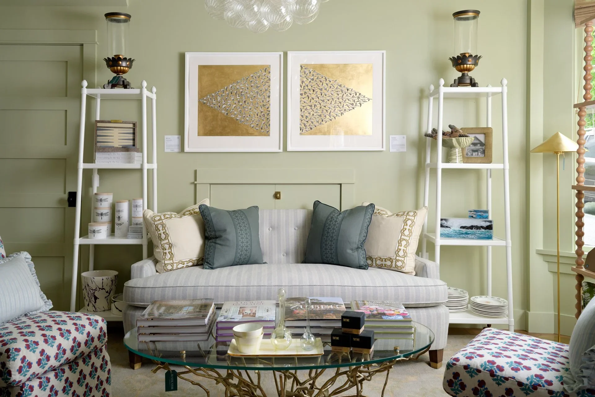 Living room with pale green walls, a white striped sofa with blue and white pillows, floral upholstered chairs, a glass coffee table with magazines and decor, wall art featuring two framed abstract prints, and white shelves displaying decorative items and books.
