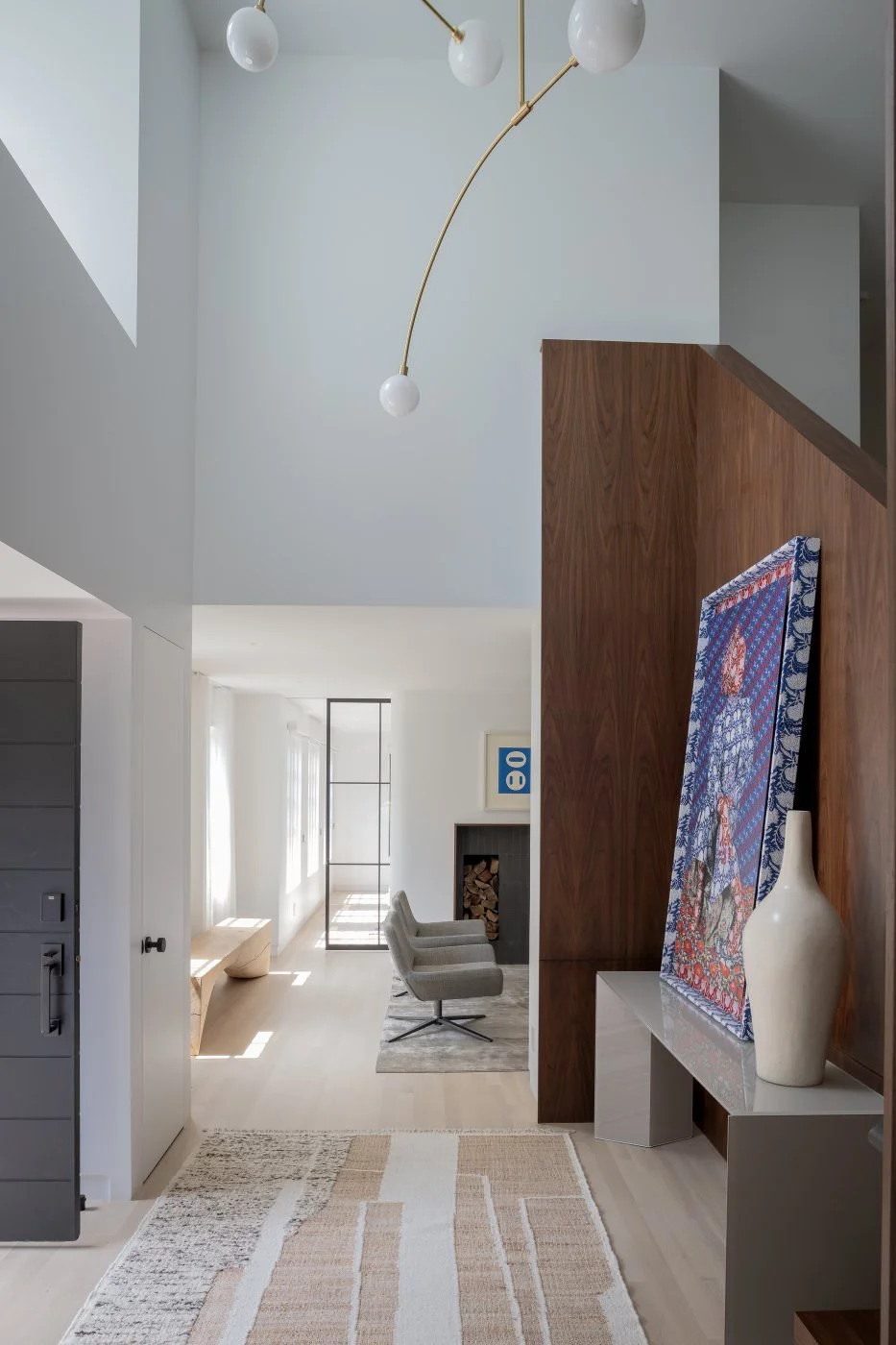 Modern living room with white walls and light wood flooring, a gray armchair, a fireplace, and a large colorful artwork on a wooden partition, with a white vase on a bench.