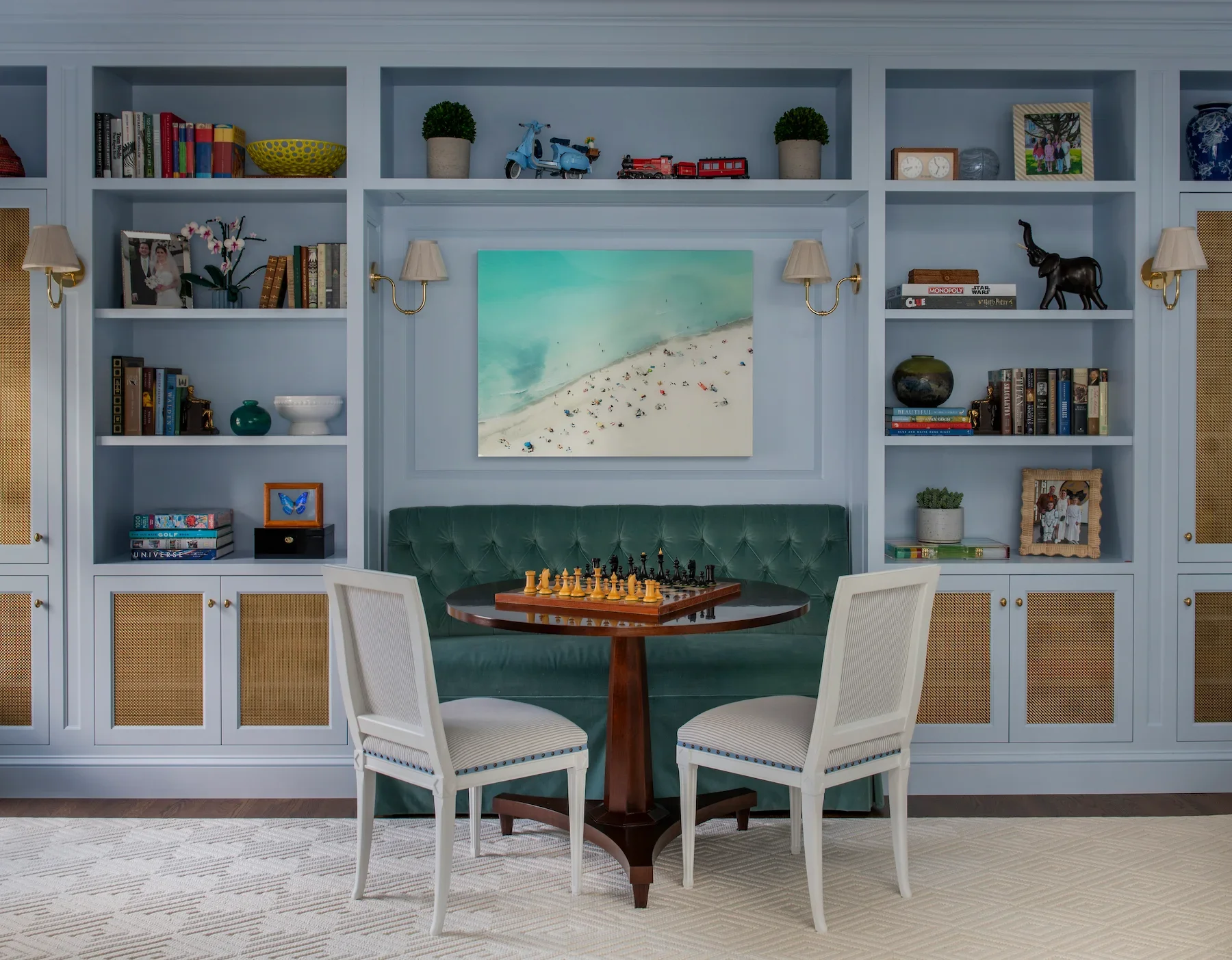 Living room with built-in white shelves filled with books, plants, decorative items, and framed photos. A large coastal painting hangs between two wall-mounted lamps. There is a green velvet tufted sofa, a round wooden table with a chessboard, and two white upholstered chairs.