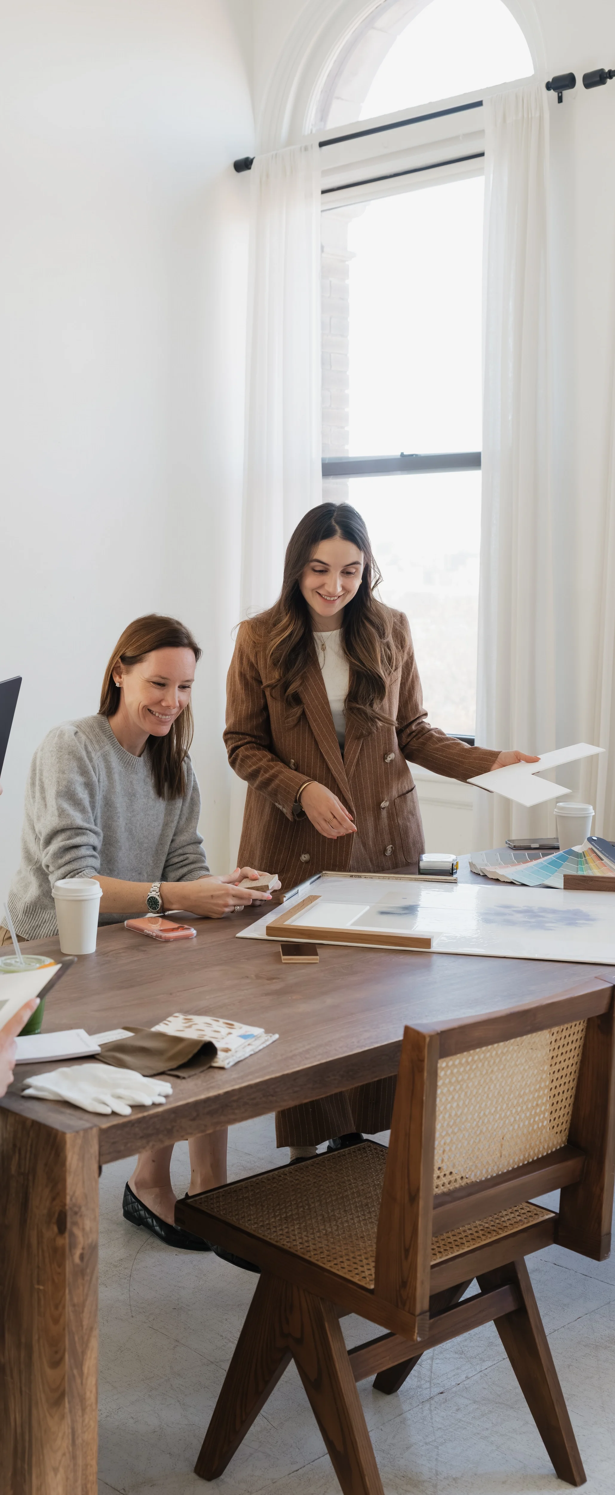 Two women are in an office, one standing and smiling while holding a piece of paper, and the other sitting, smiling and holding a color swatch book. The office has a large window with white curtains, a wooden table with various items, and wooden chairs.