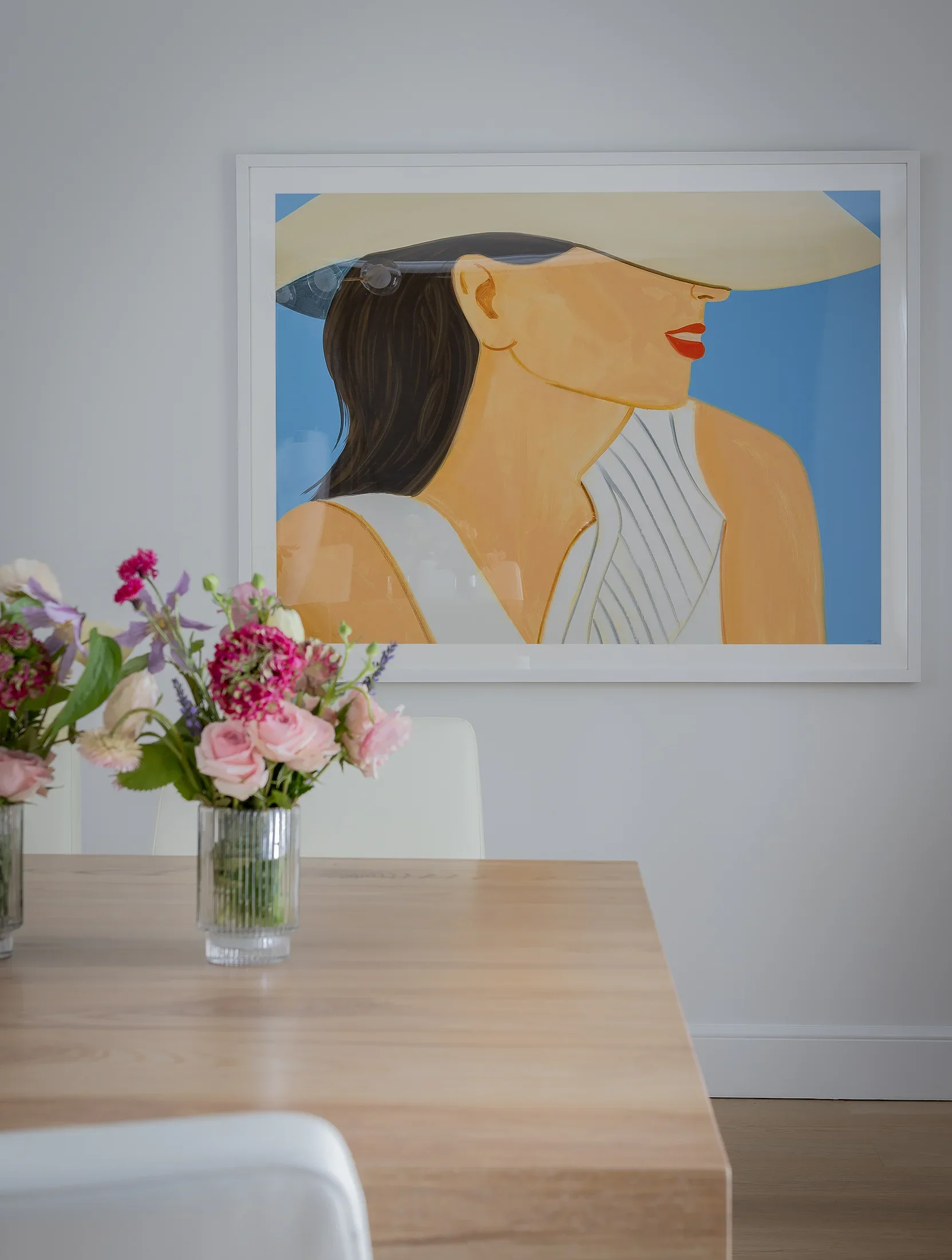A wooden table with a glass vase of pink, purple, and white flowers, with a large framed artwork of a woman wearing a wide-brimmed hat on the wall behind.