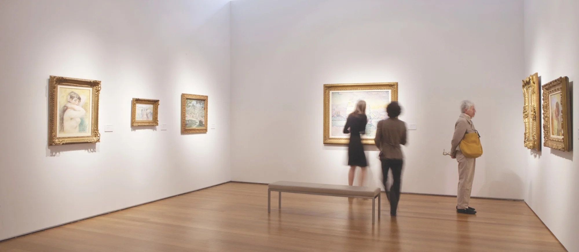 People viewing framed artwork in a gallery with white walls and wooden floors.