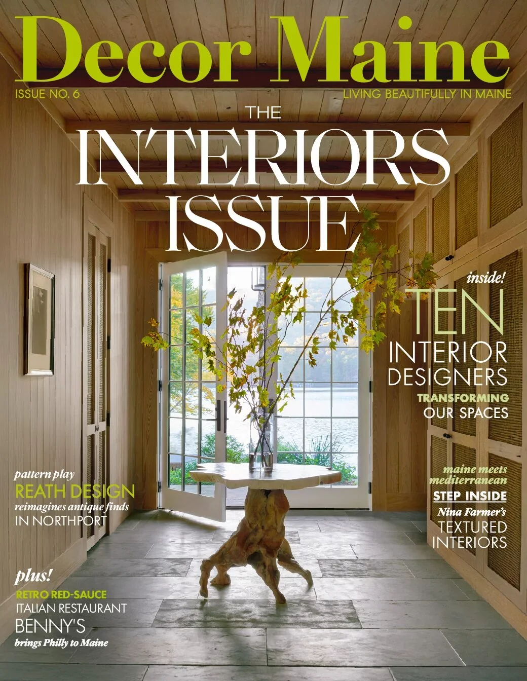 Cover of Decor Maine magazine featuring a wooden interior with a large glass door opening to an outdoor view of water and trees. A unique table with a twisted tree trunk base and a vase with leafy branches is in the center.